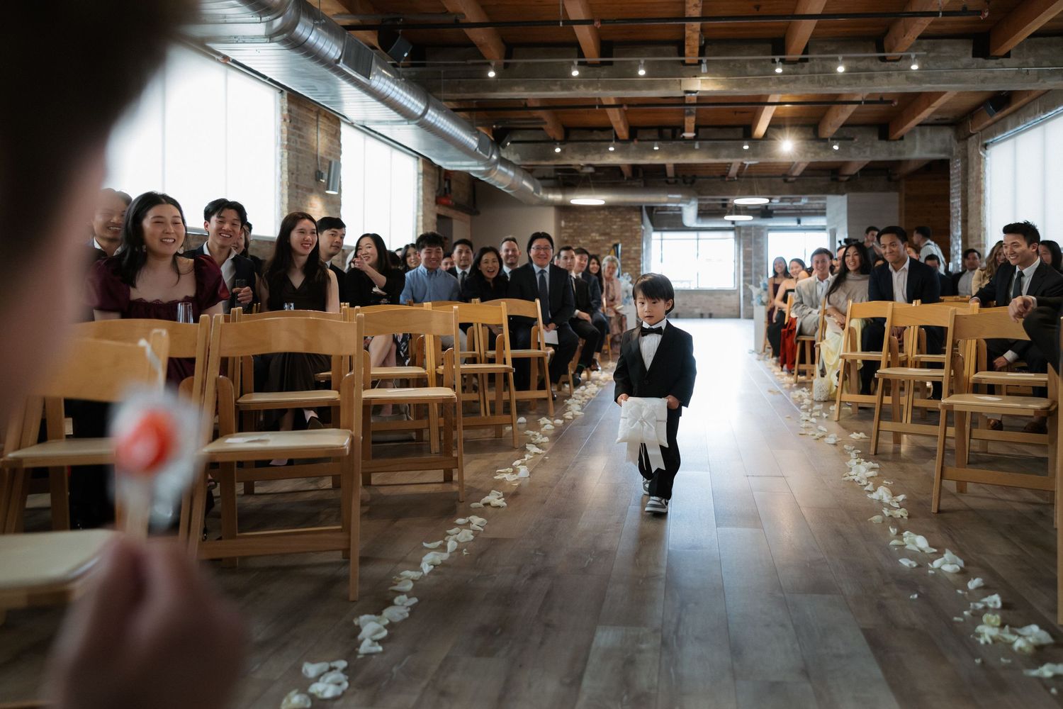 A young ring bearer walks down the aisle at an elegant indoor wedding ceremony with guests seated on wooden chairs.
