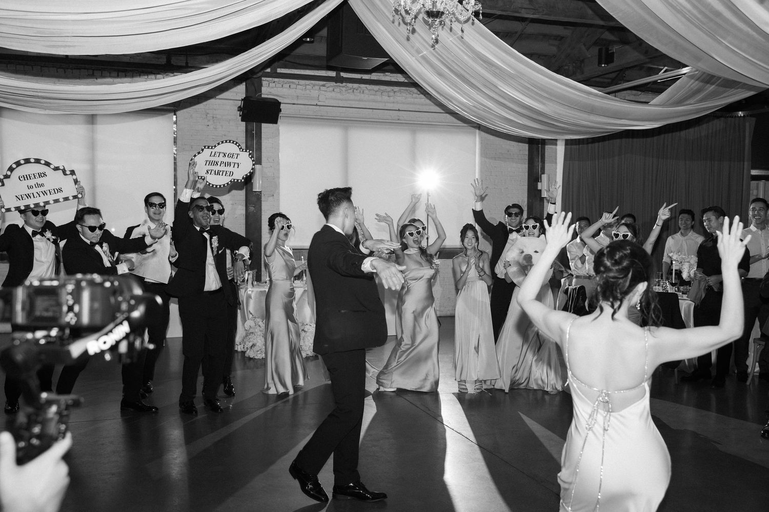 Black and white photo of bride and groom dancing with cheering guests at lively wedding reception.