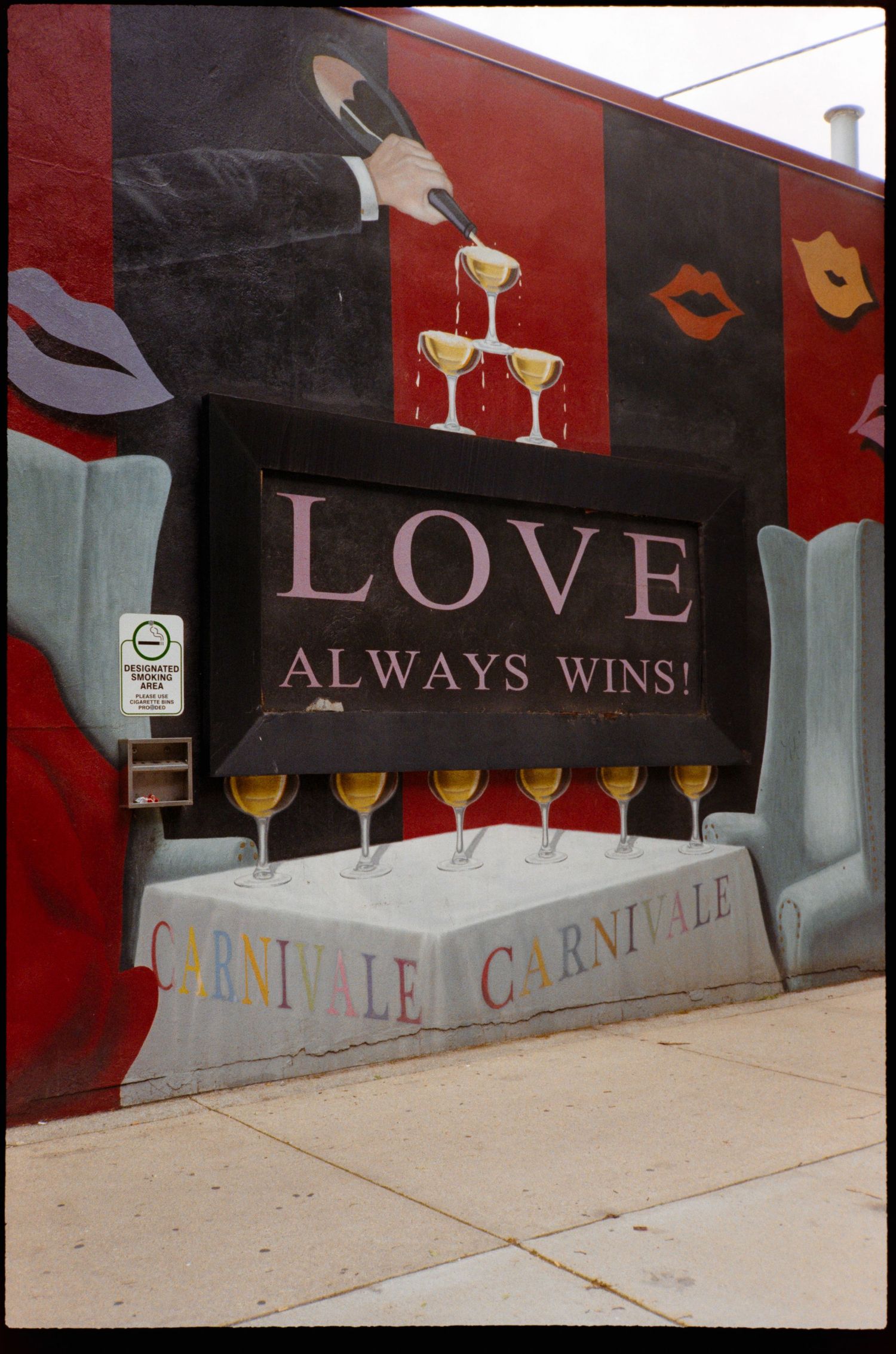 Colorful mural on a building wall reading Love Always Wins with champagne glasses and Carnivale text on sidewalk.