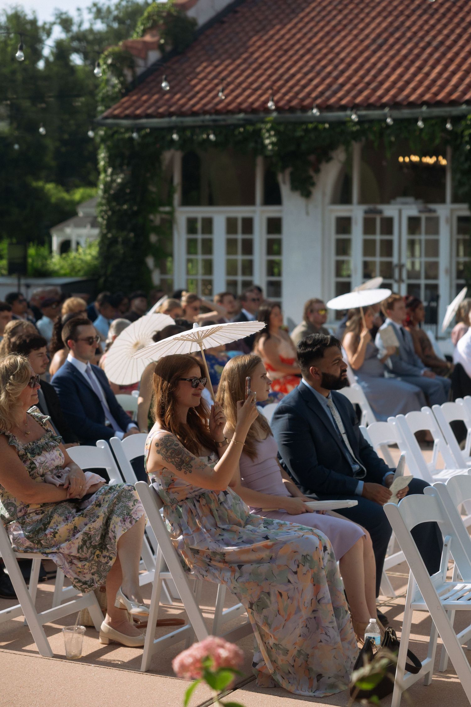 Outdoor wedding ceremony guests seated in white chairs, some holding parasols in the summer sun.