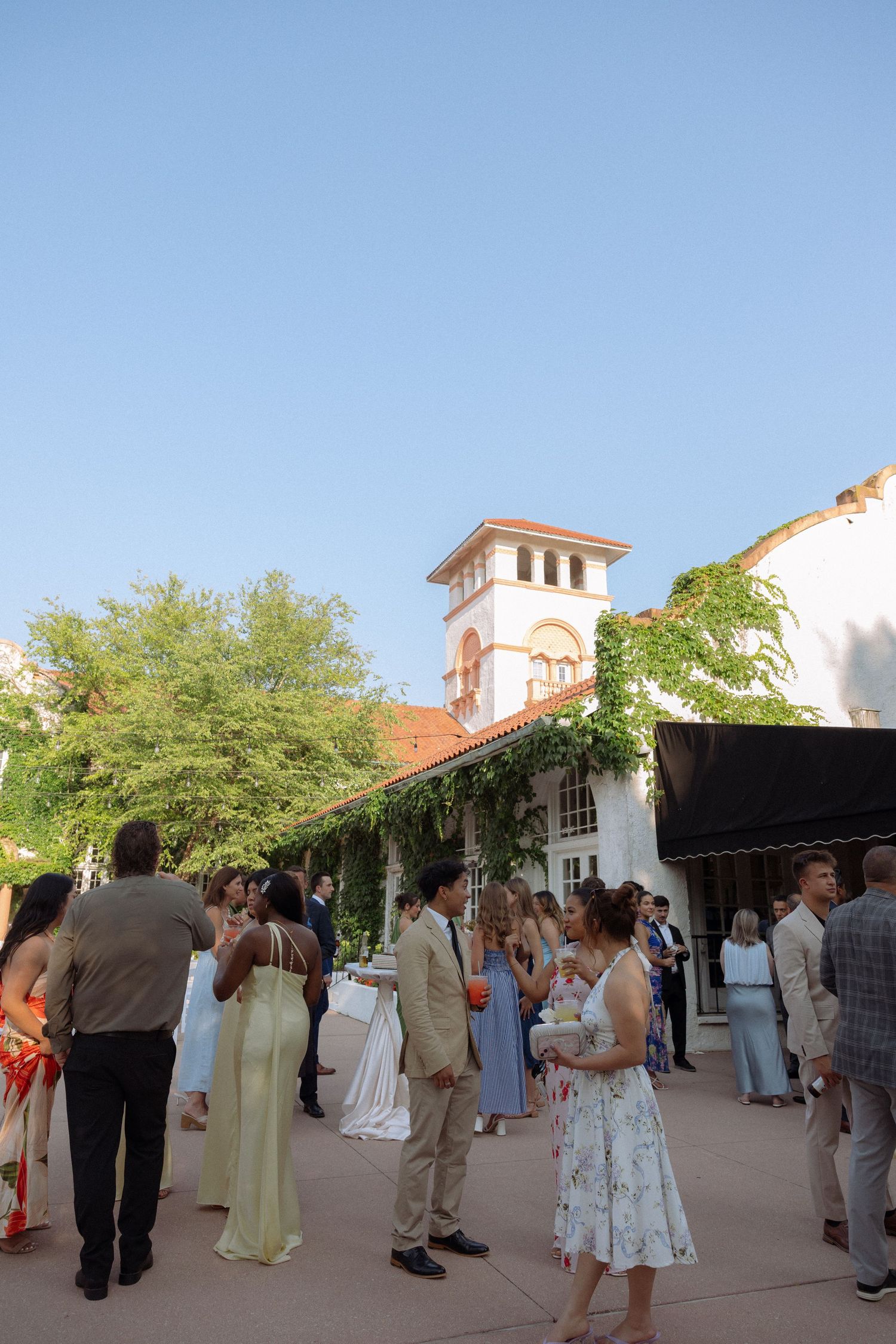 Guests mingle outdoors at an elegant courtyard wedding reception near a historic building with a bell tower.