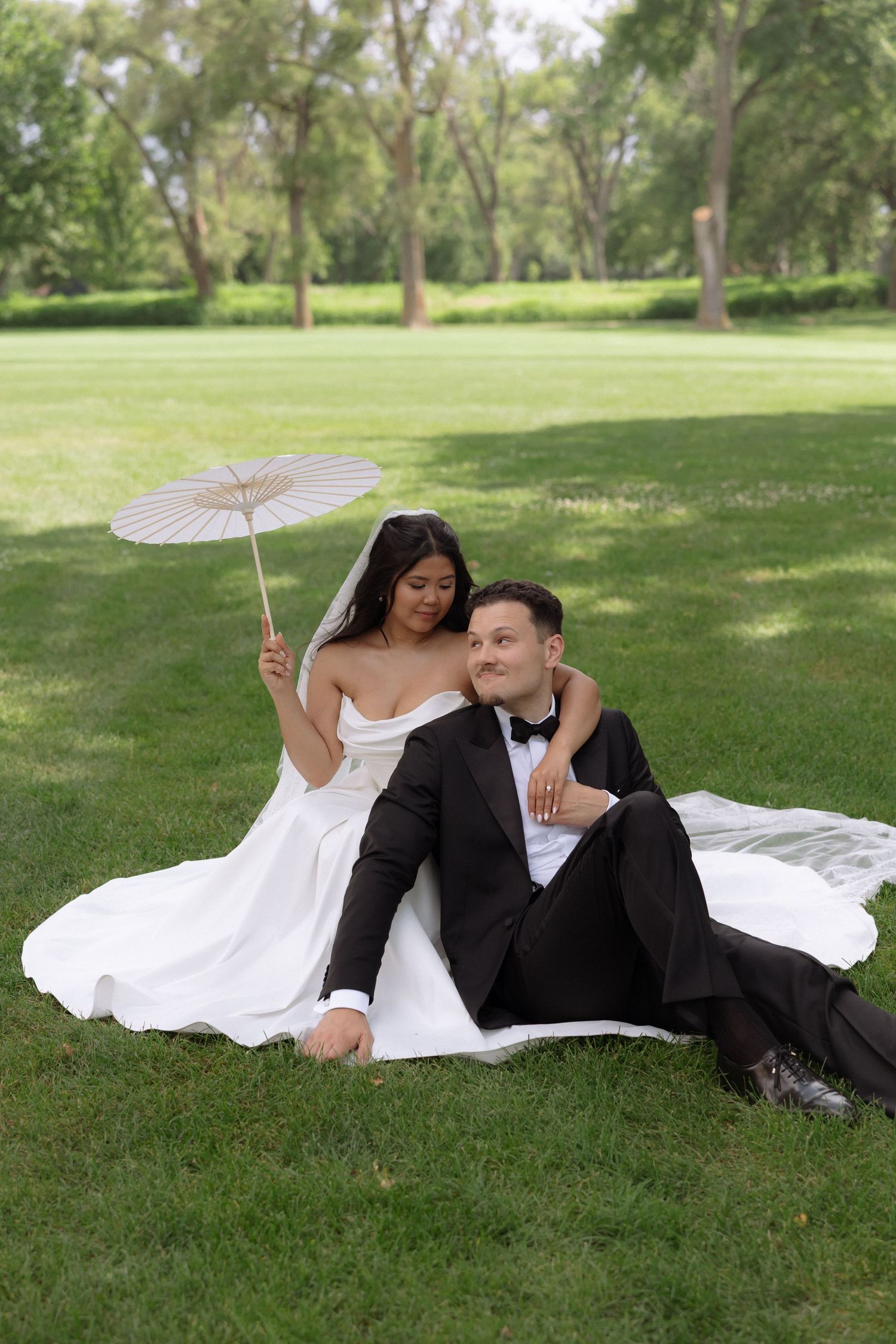 Bride and groom sitting on grass in a park, bride holding a white parasol umbrella on their wedding day.