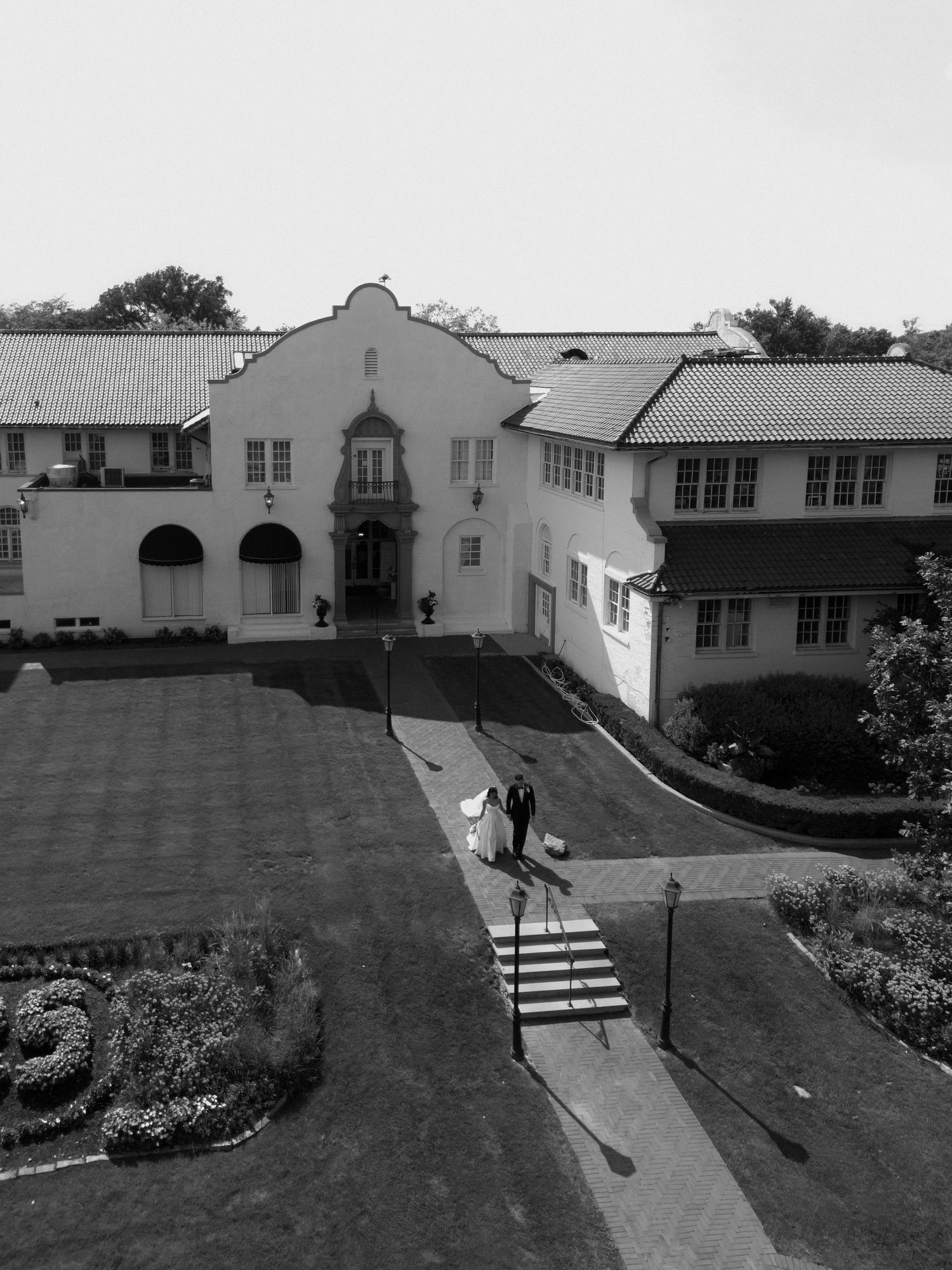 Black and white aerial view of bride and groom walking outside an elegant estate venue.