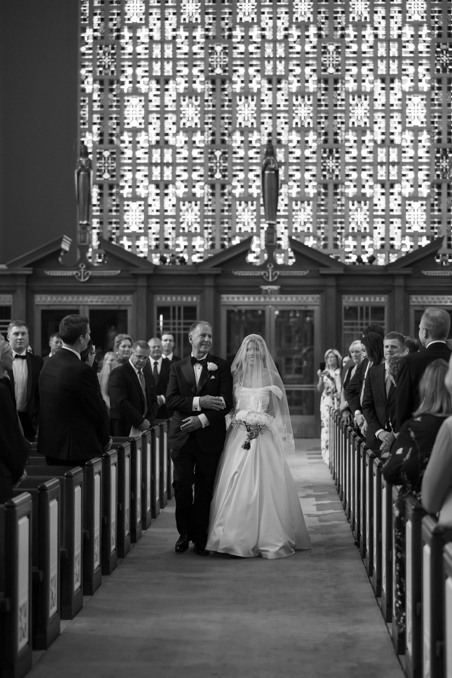 Black and white photo of bride walking down church aisle with father during wedding ceremony.
