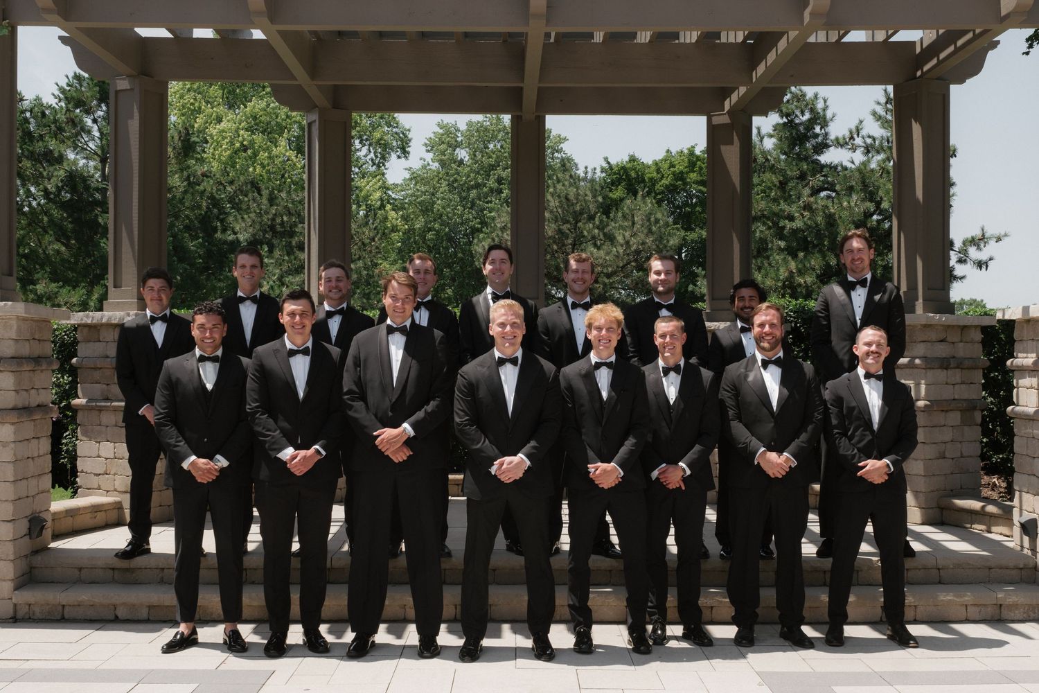 Groomsmen in black tuxedos pose together outdoors under a pergola for a formal wedding party portrait.