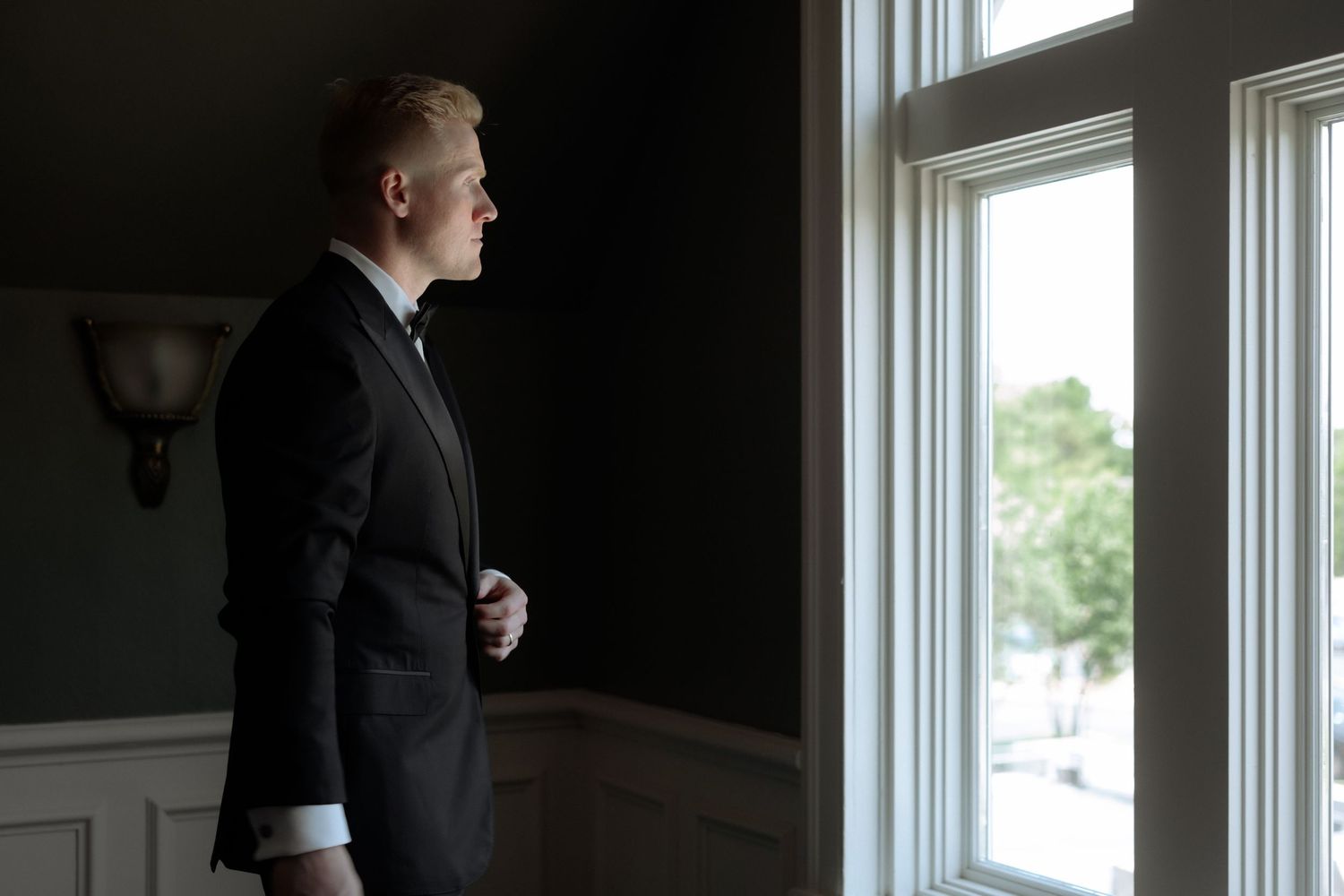 Man in black tuxedo gazing thoughtfully out a window in an elegant dark room.