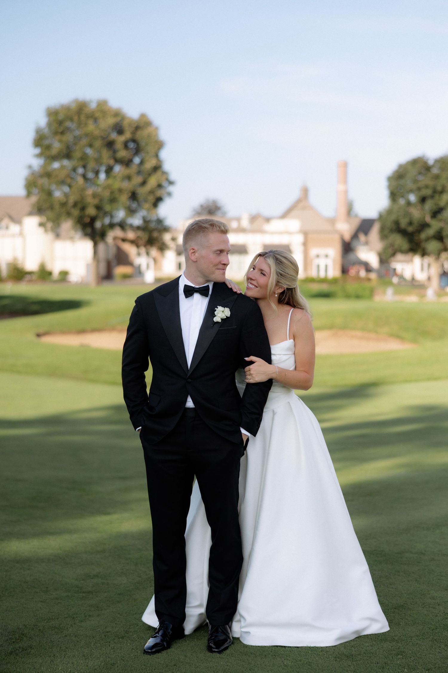 Elegant bride and groom posing together on golf course with grand estate building in background.