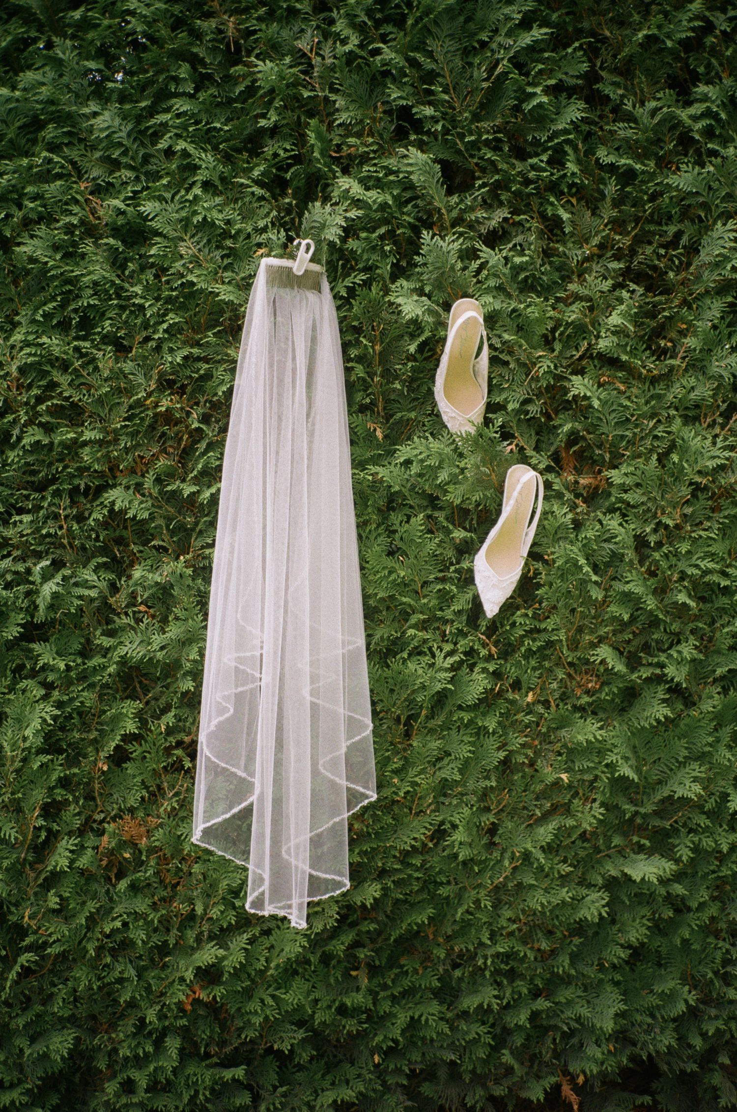 Bridal veil hanging with white heels against lush green hedge backdrop for wedding day details.