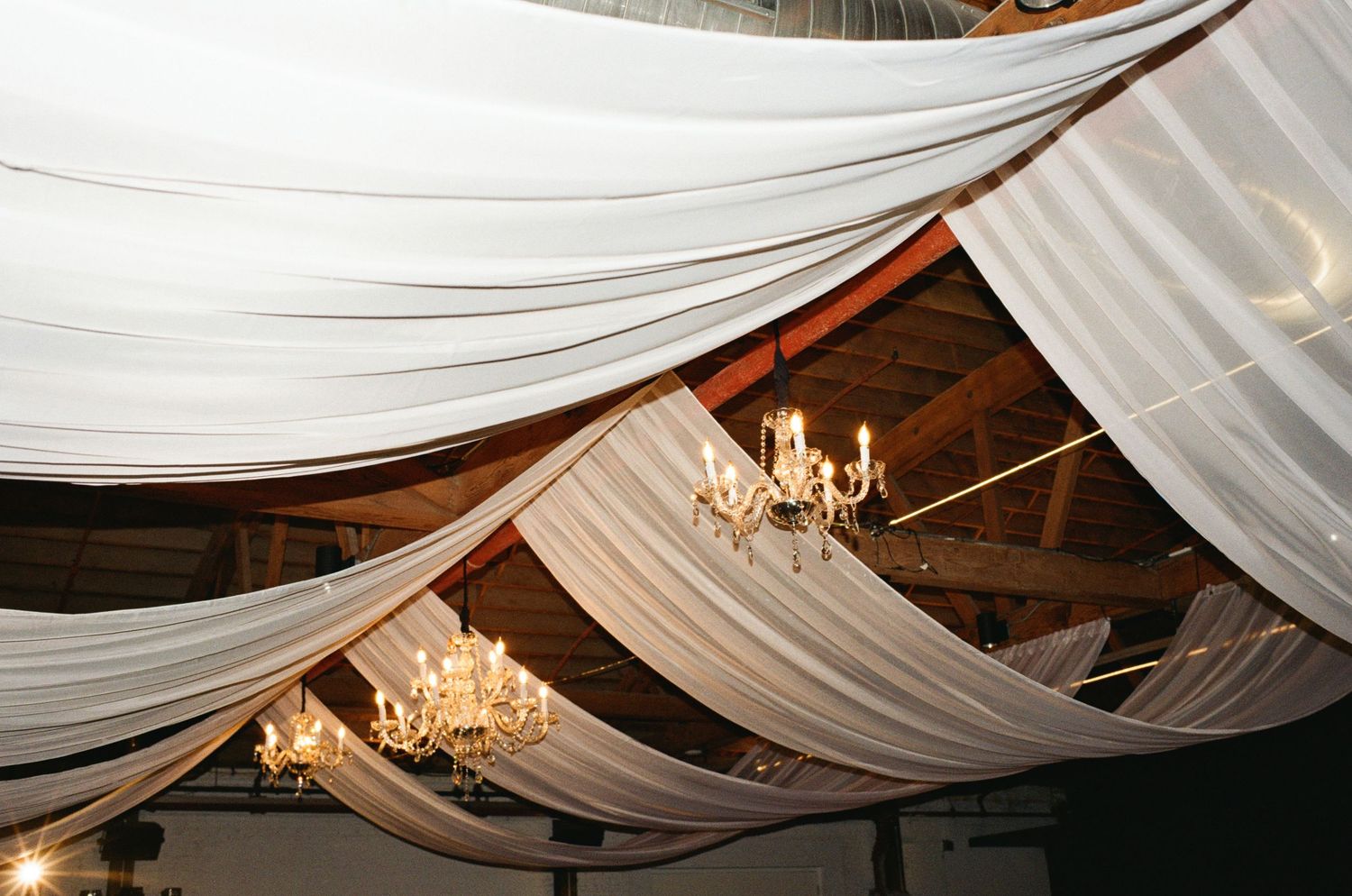 Elegant white fabric draping on barn ceiling with crystal chandeliers at a rustic wedding venue.