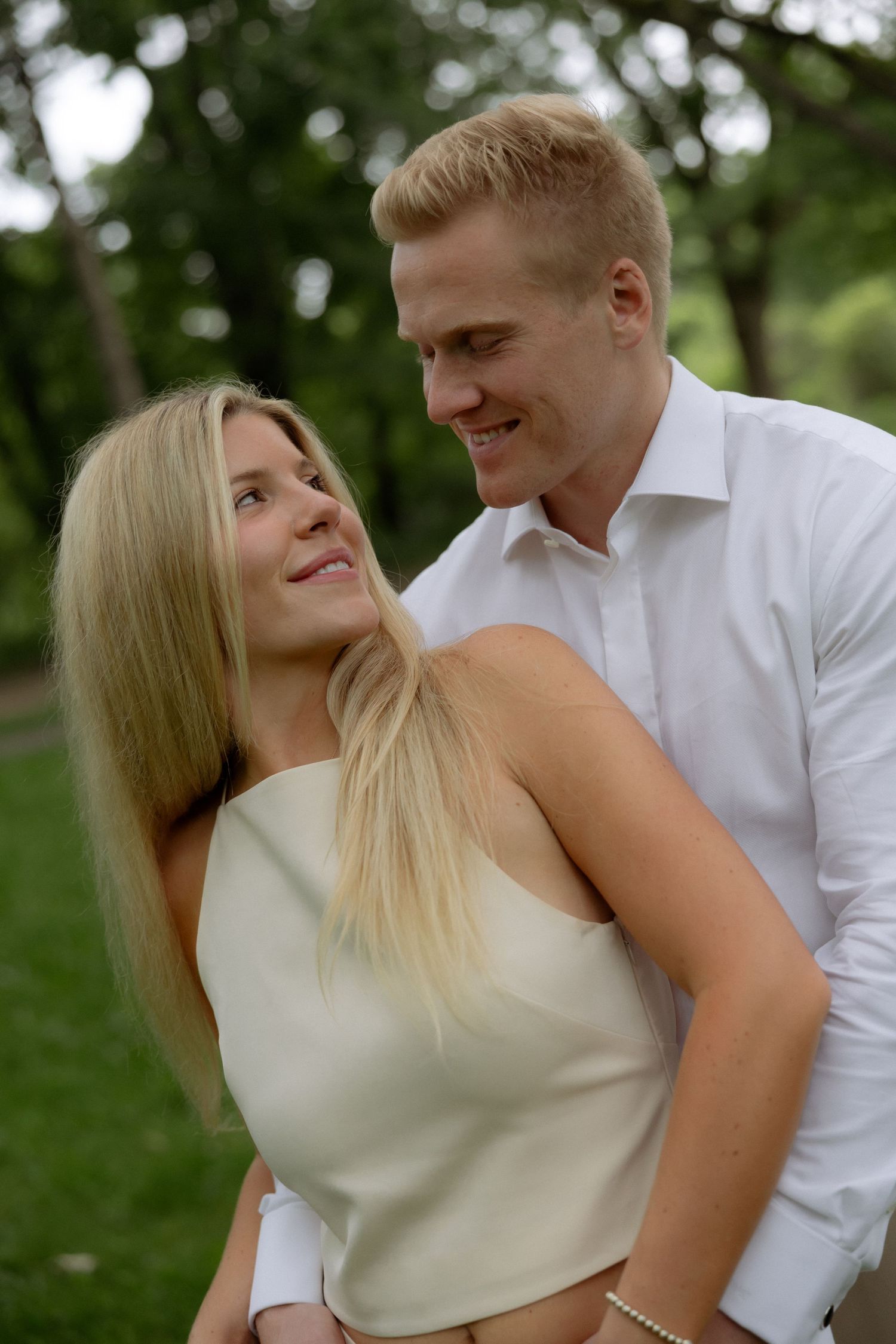 A smiling blonde woman and tall man in a white shirt embrace warmly in a lush green park.