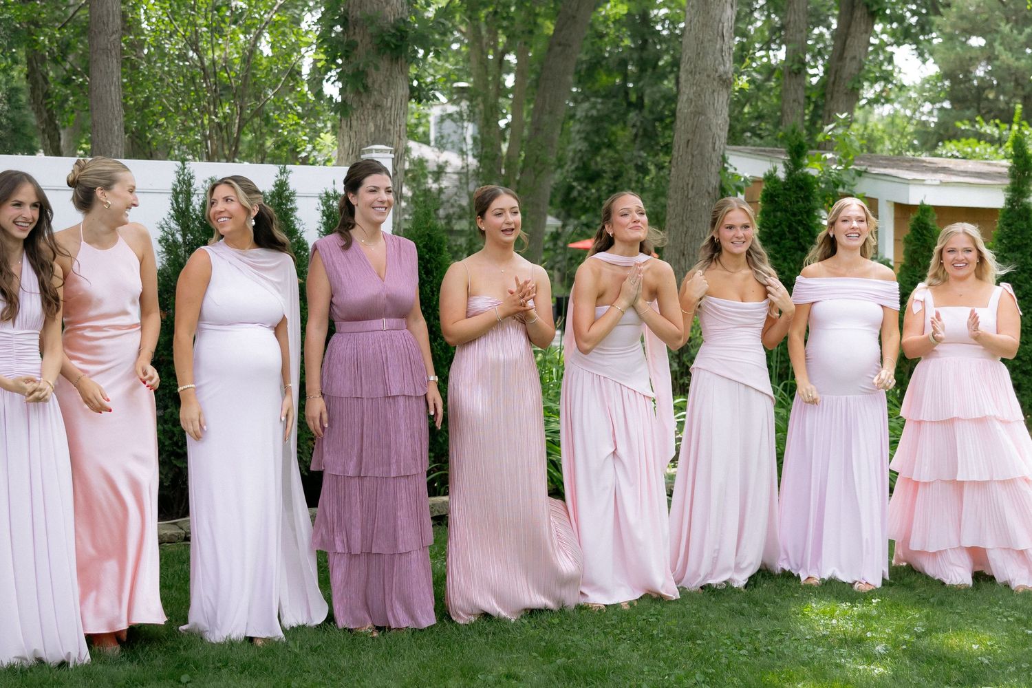 Nine bridesmaids in mismatched pink dresses laughing together outdoors at a garden wedding celebration.