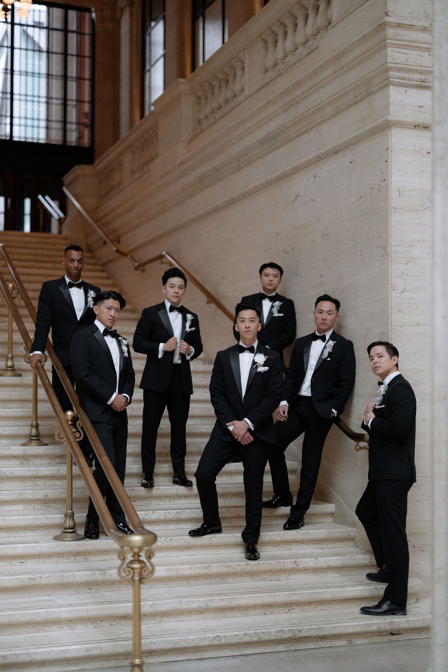 Seven groomsmen in black tuxedos pose on ornate marble staircase with gold railing.
