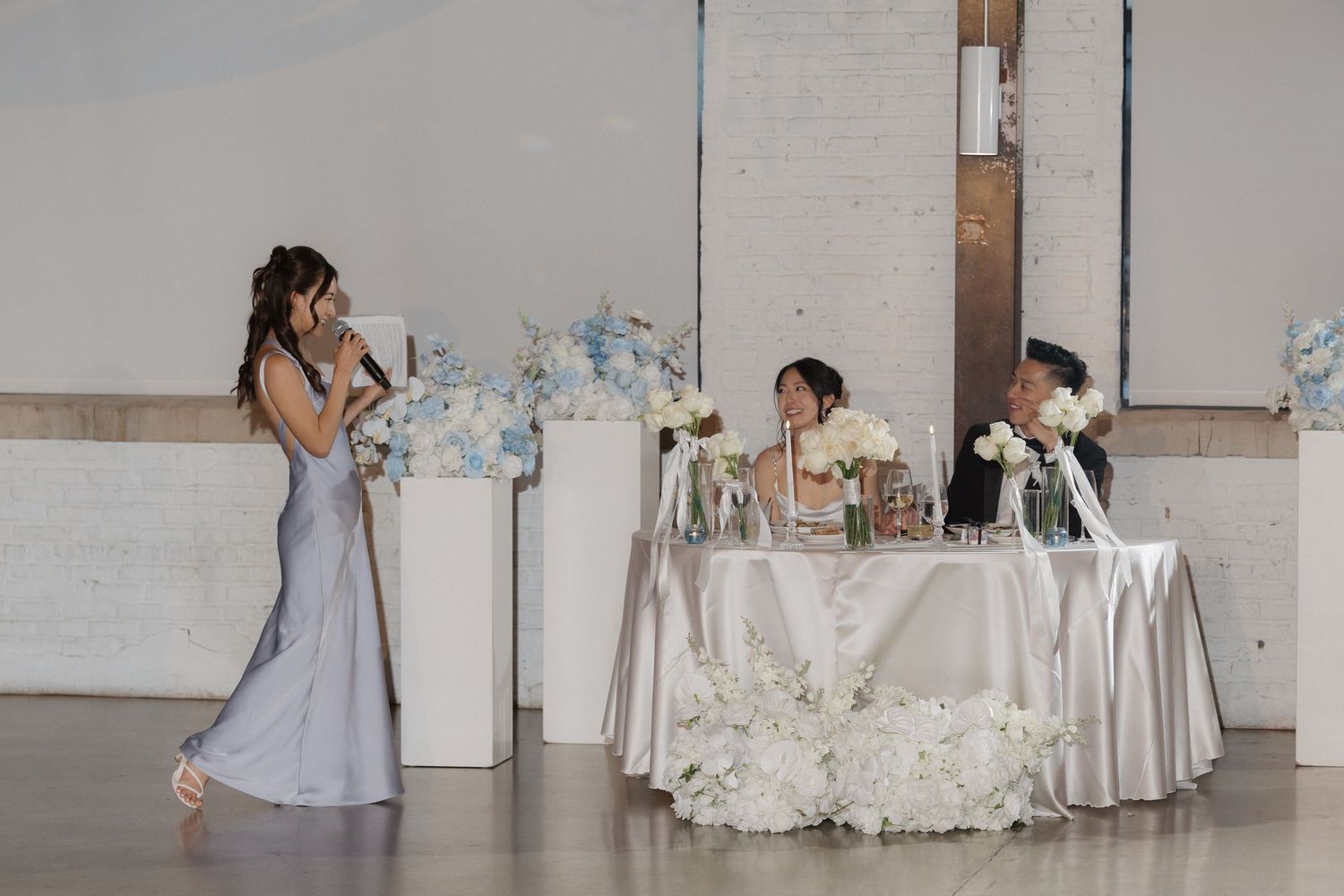 Woman in silver gown giving speech at elegant wedding reception with blue floral decorations.
