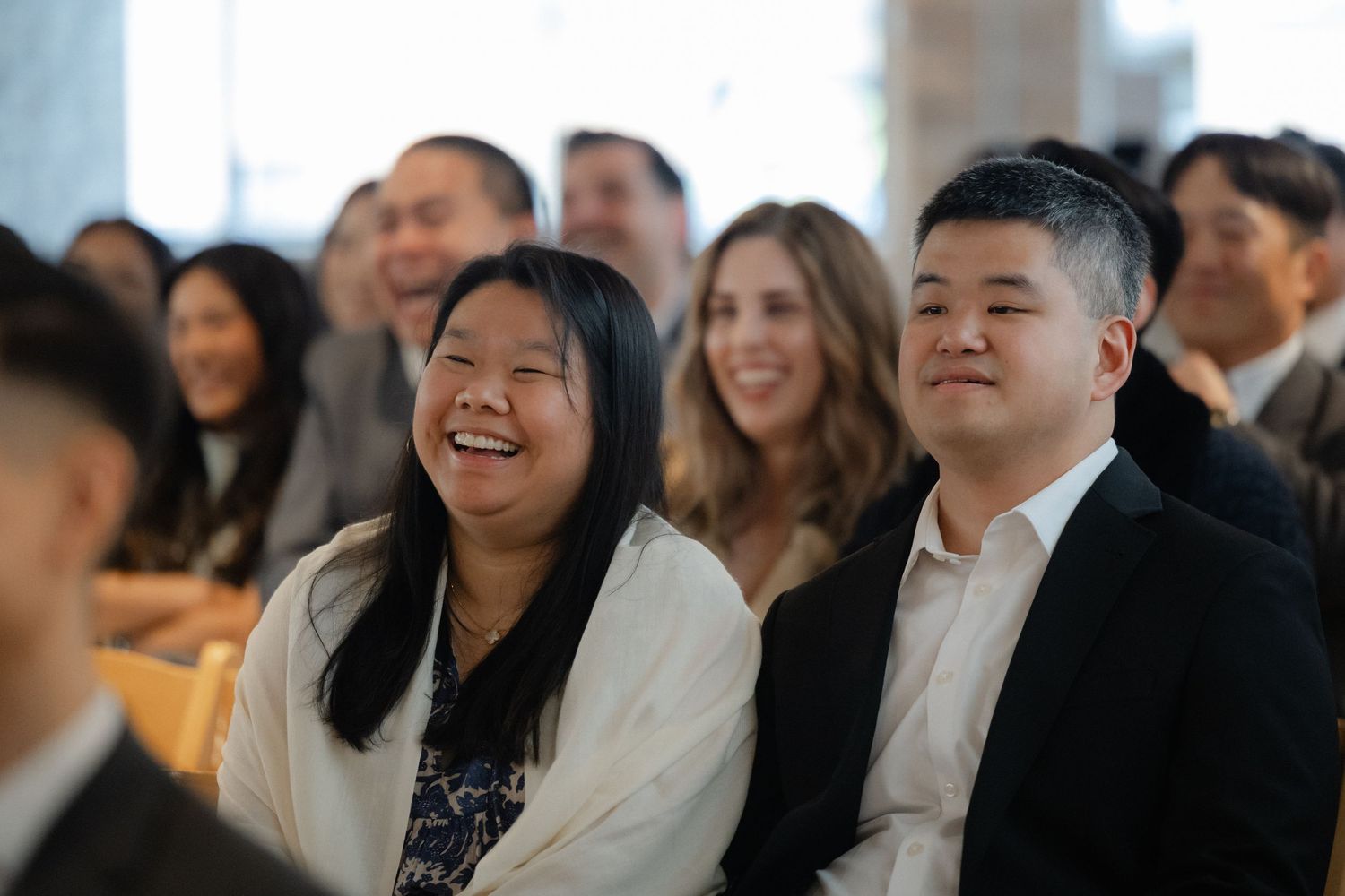 Two smiling Asian attendees laugh together at an indoor event or ceremony, seated among a diverse crowd.