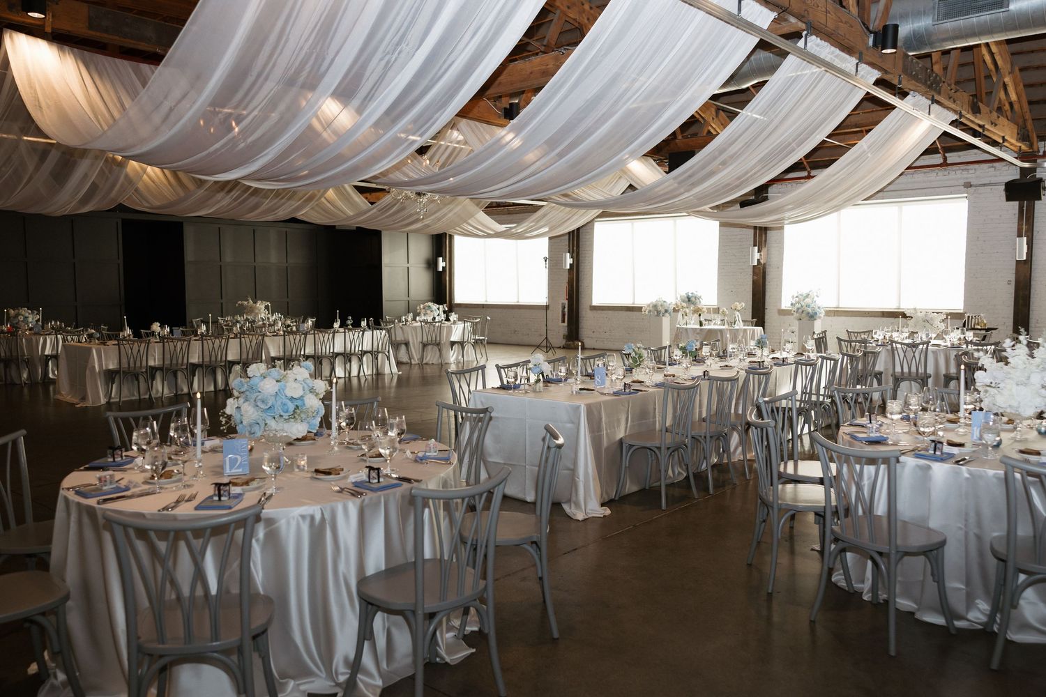 Elegant wedding reception hall with white draped ceiling, silver chiavari chairs, and round tables with white linens.