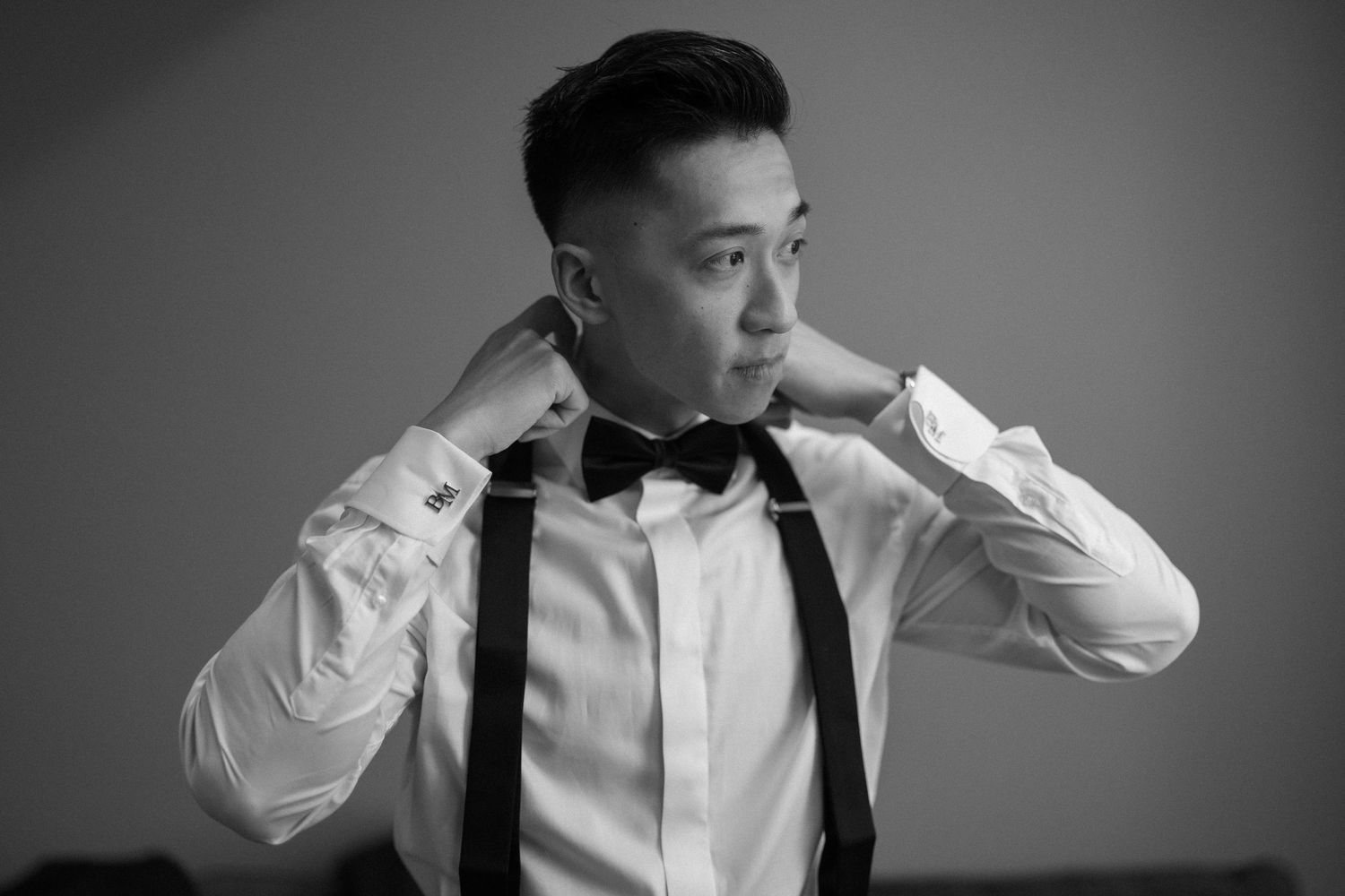 Black and white photo of a young Asian man adjusting his bow tie, wearing a white dress shirt with suspenders.
