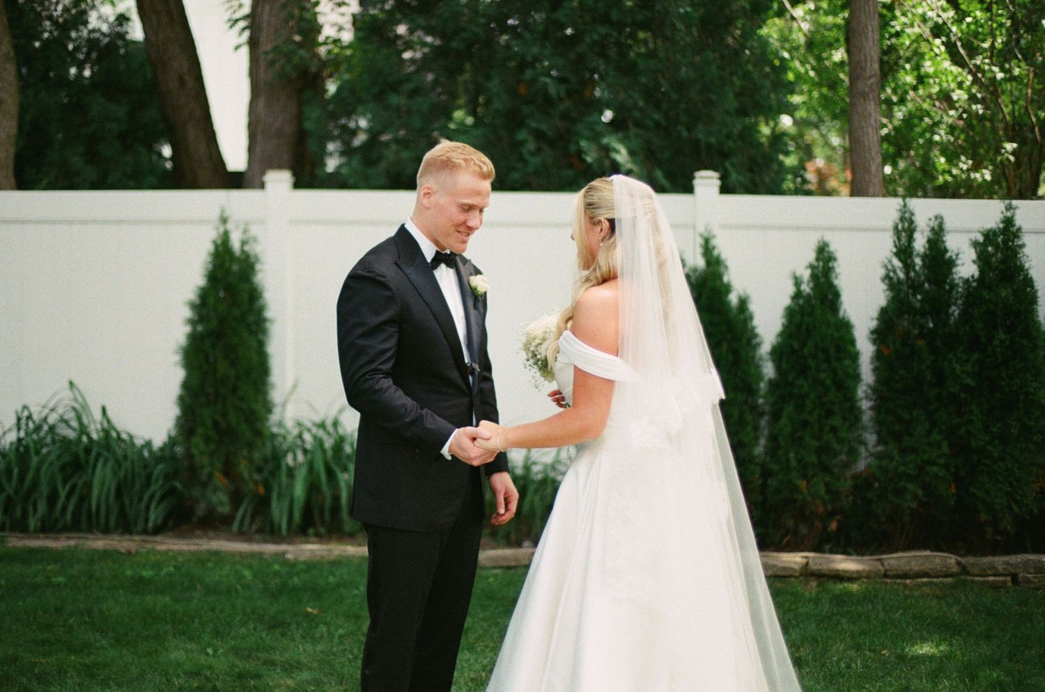 Bride and groom share a tender first look moment outdoors, with lush green hedges and white fence backdrop.