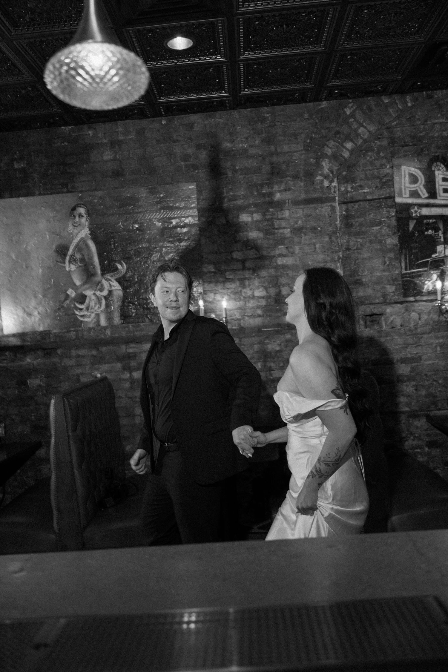 Black and white photo of newlyweds holding hands and laughing in a rustic brick-walled venue.