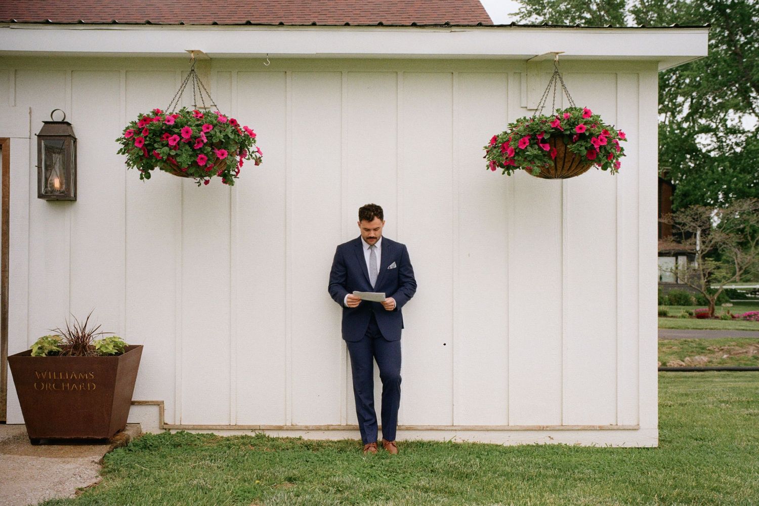 Groom in navy suit reads vows outside white barn adorned with hanging pink flower baskets at outdoor wedding venue.