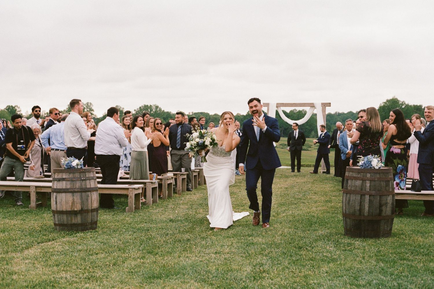 Bride and groom walk down outdoor wedding aisle lined with wooden benches and barrel decorations on a green lawn.