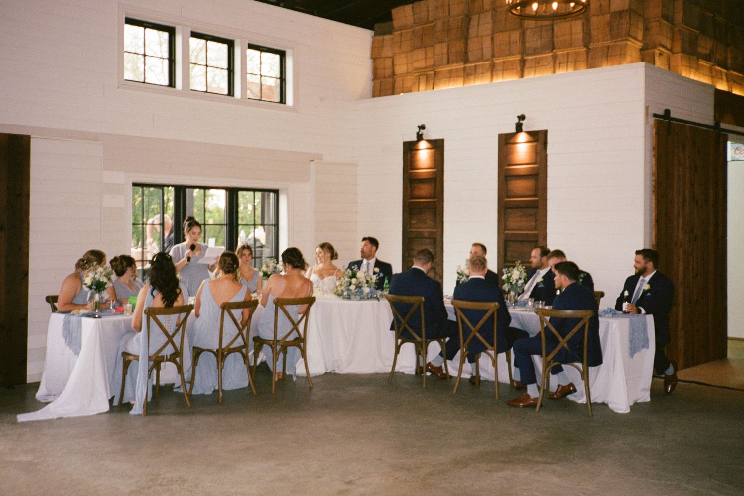 Wedding party and guests seated at elegantly decorated tables in a rustic venue.