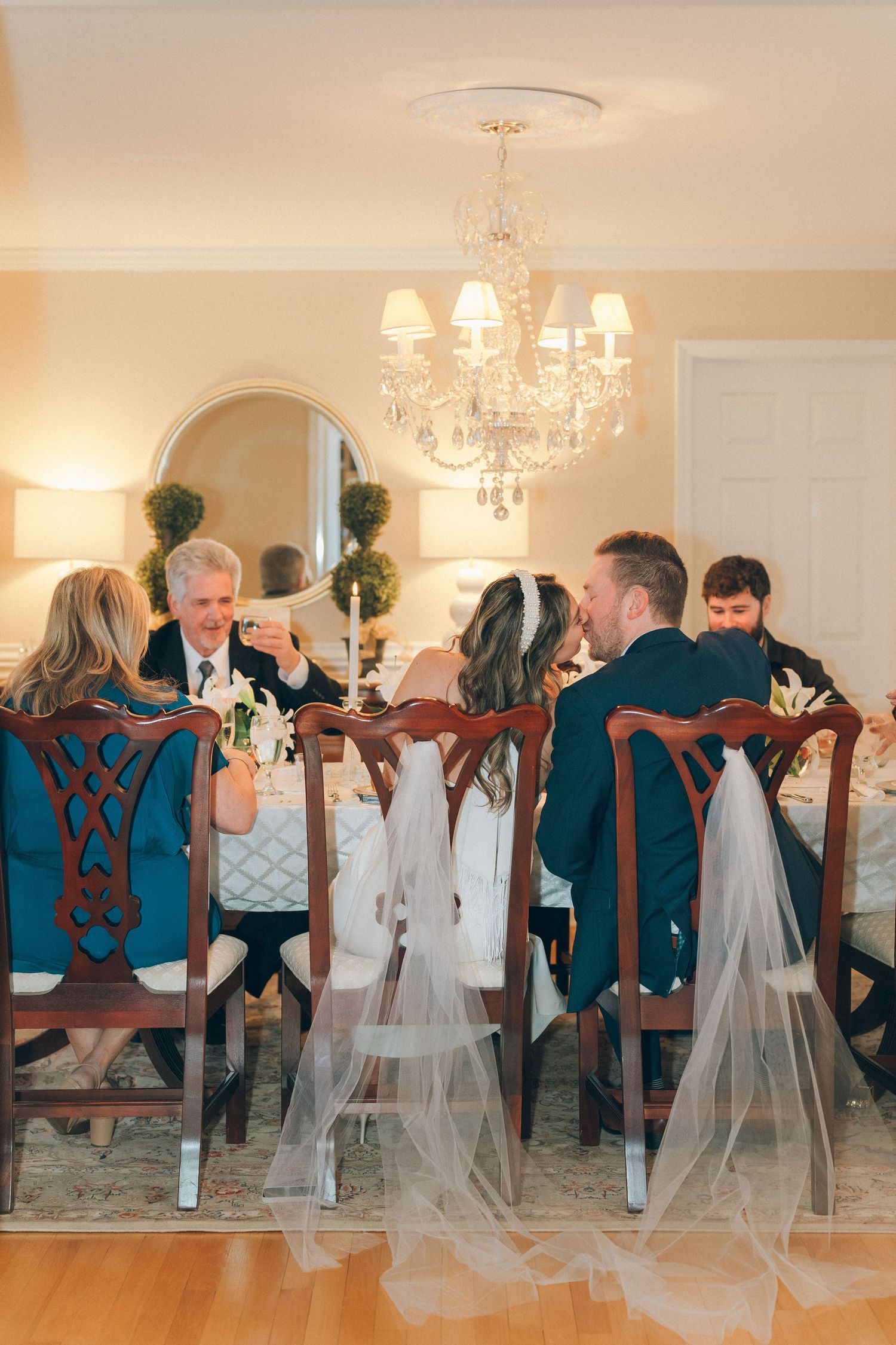 Bride with veil and groom seated with family at elegant candlelit wedding dinner under a crystal chandelier.