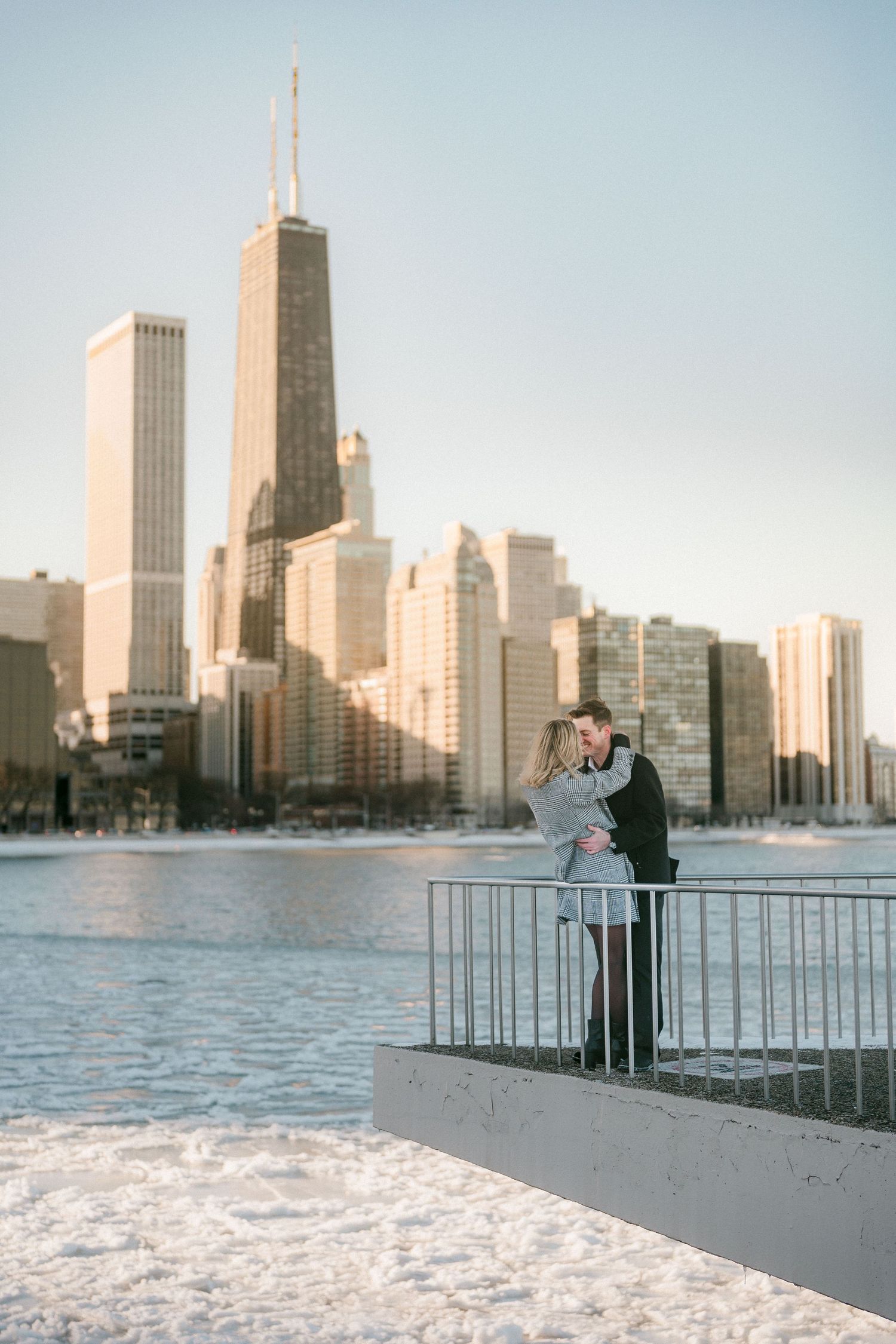 Couple kissing near railing with Chicago skyline and frozen lake visible in the background on sunny winter day.