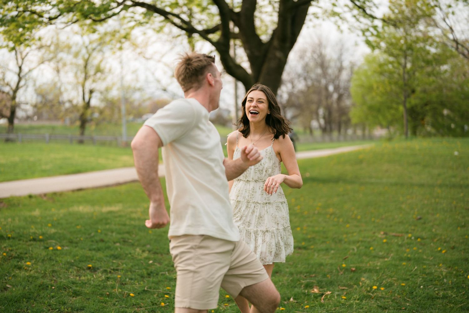 A laughing couple playfully chases each other across a green park lawn in spring.