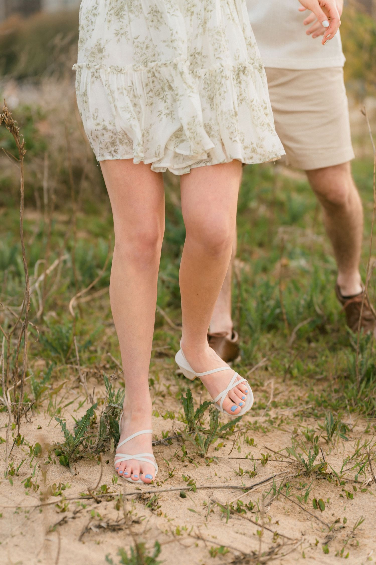 Close-up of woman in floral dress and white sandals walking on sandy path.