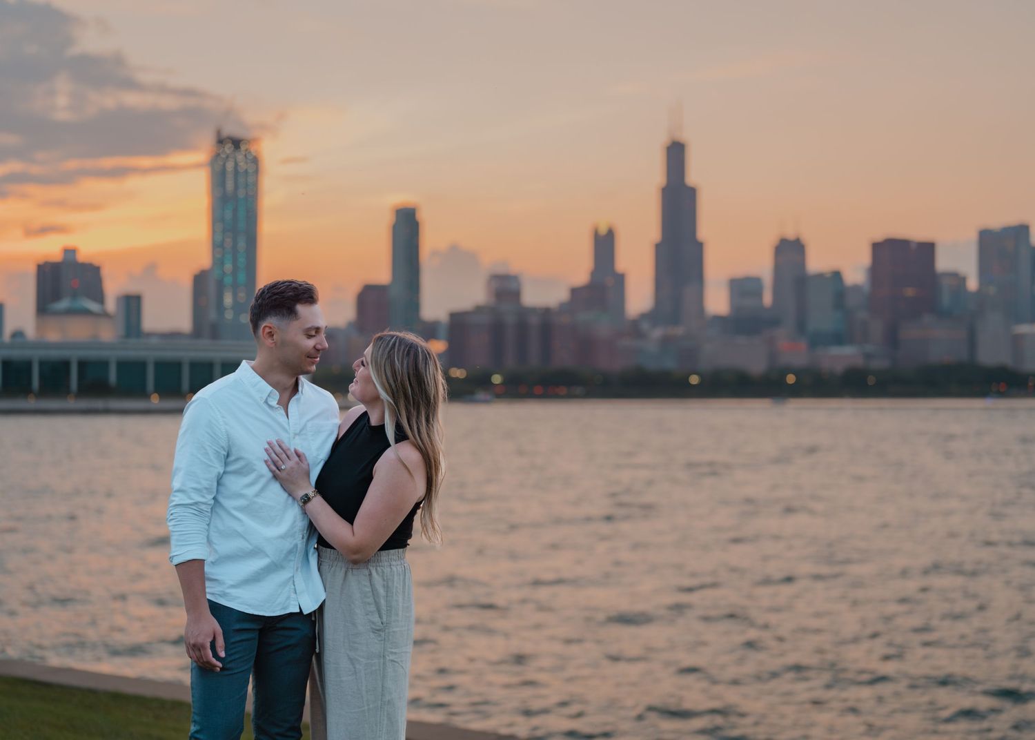 Engaged couple gazing at each other by a lake with a vibrant Chicago sunset skyline.