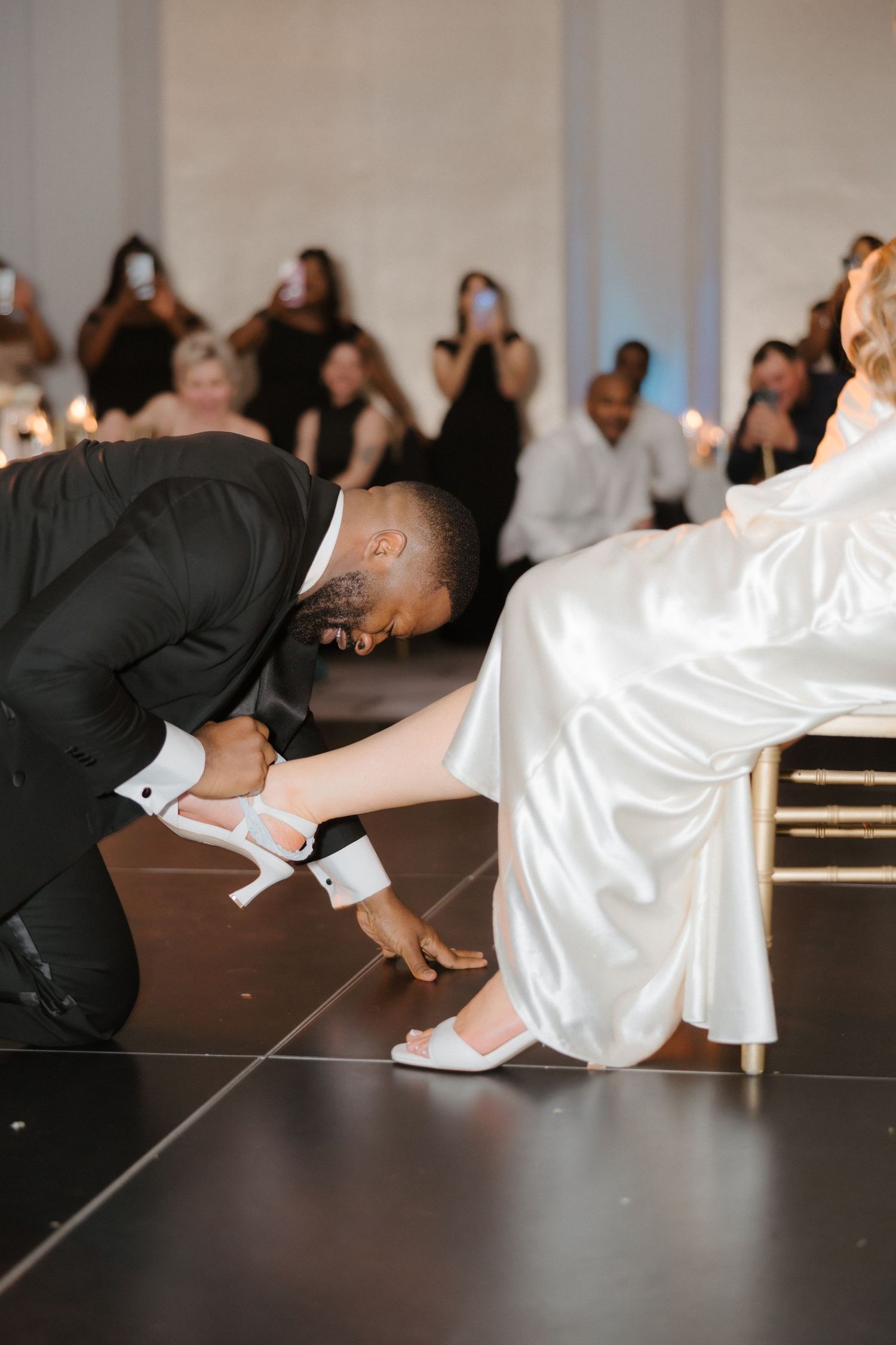 Groom kneeling to place white heels on bride seated in chair during wedding reception on dance floor.