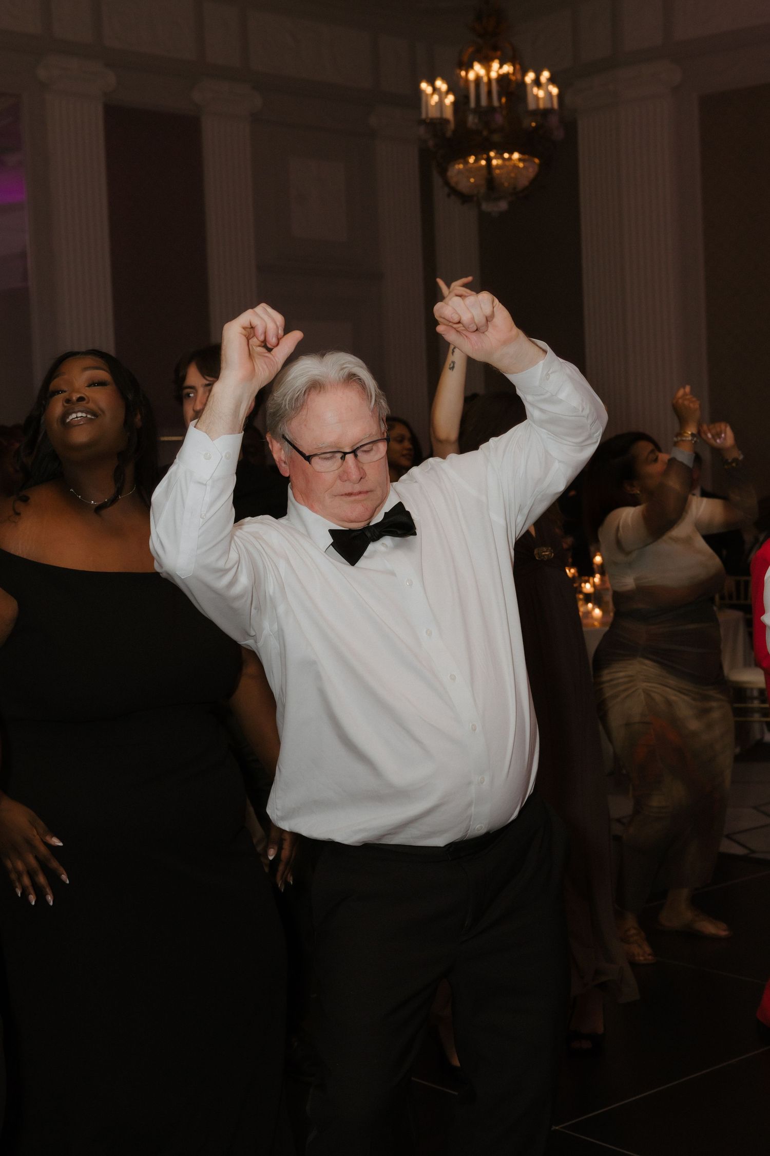 Man in bow tie dancing with arms raised at formal event under chandelier.