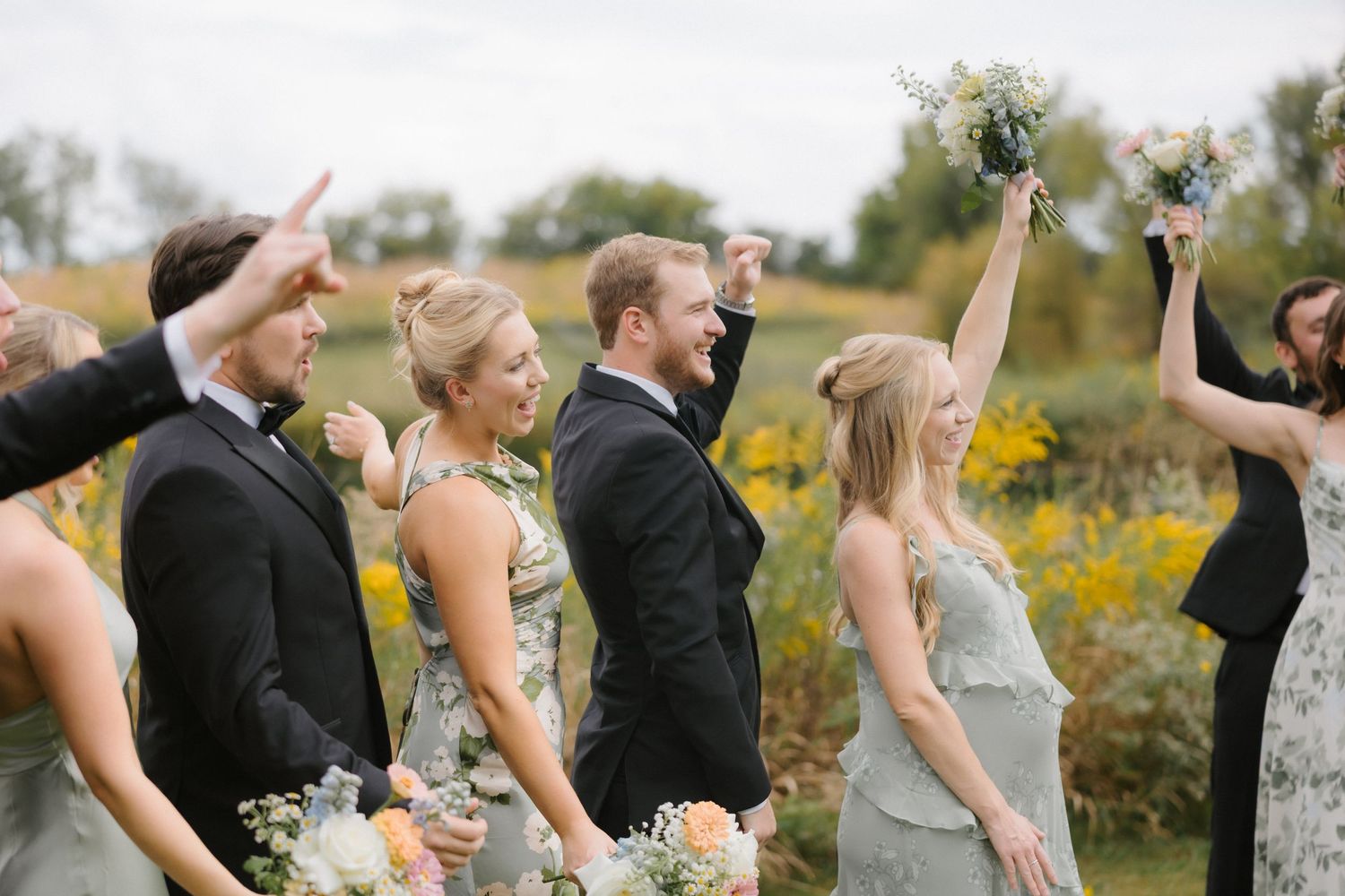 Excited wedding party members laugh and raise bouquets while celebrating outdoors near vibrant yellow wildflower fields.
