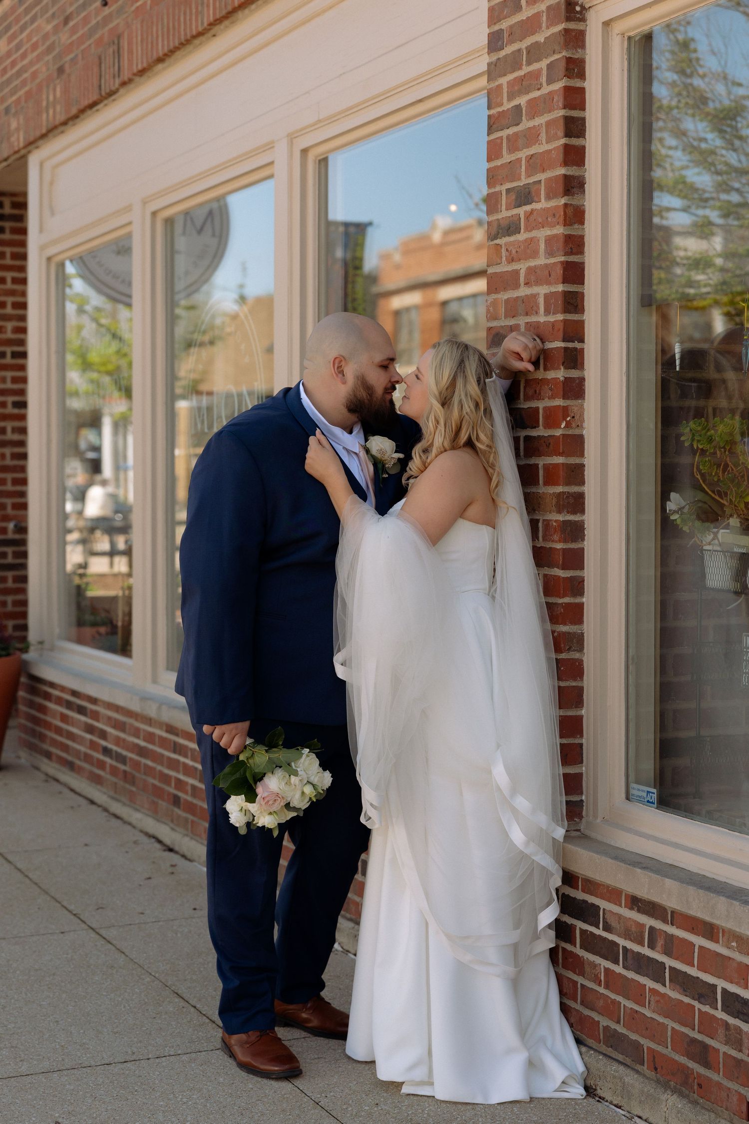 Bride and groom share a romantic kiss against a brick building exterior, bride in white gown holding bouquet.