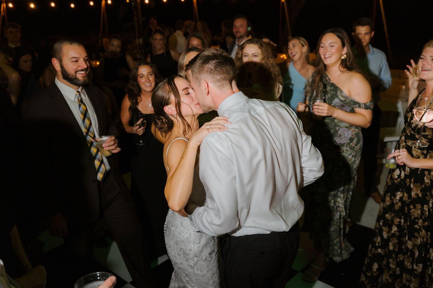 Bride and groom share a romantic kiss on the dance floor surrounded by cheering wedding reception guests.