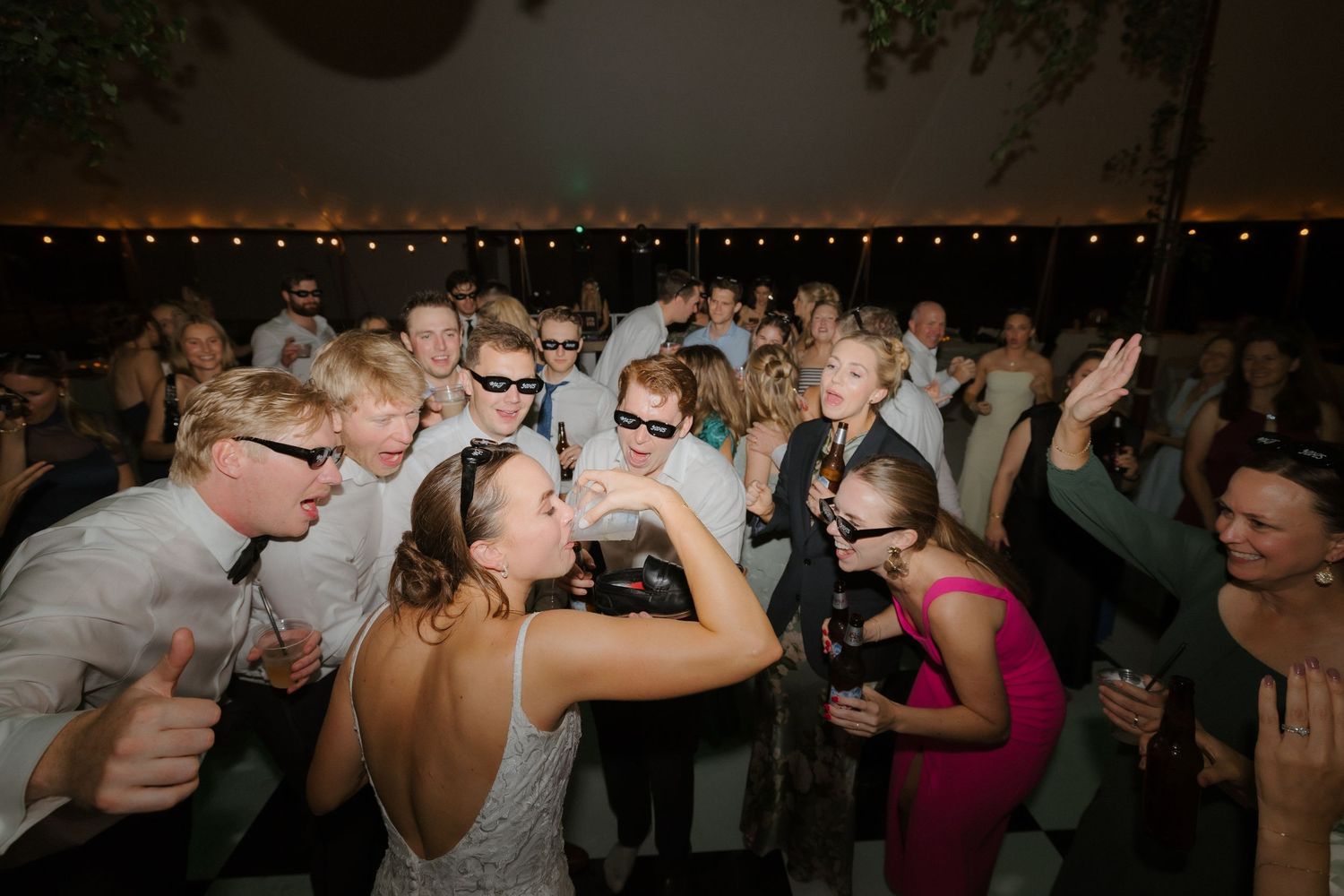 Wedding guests celebrate on the dance floor wearing sunglasses at a festive outdoor reception under string lights.