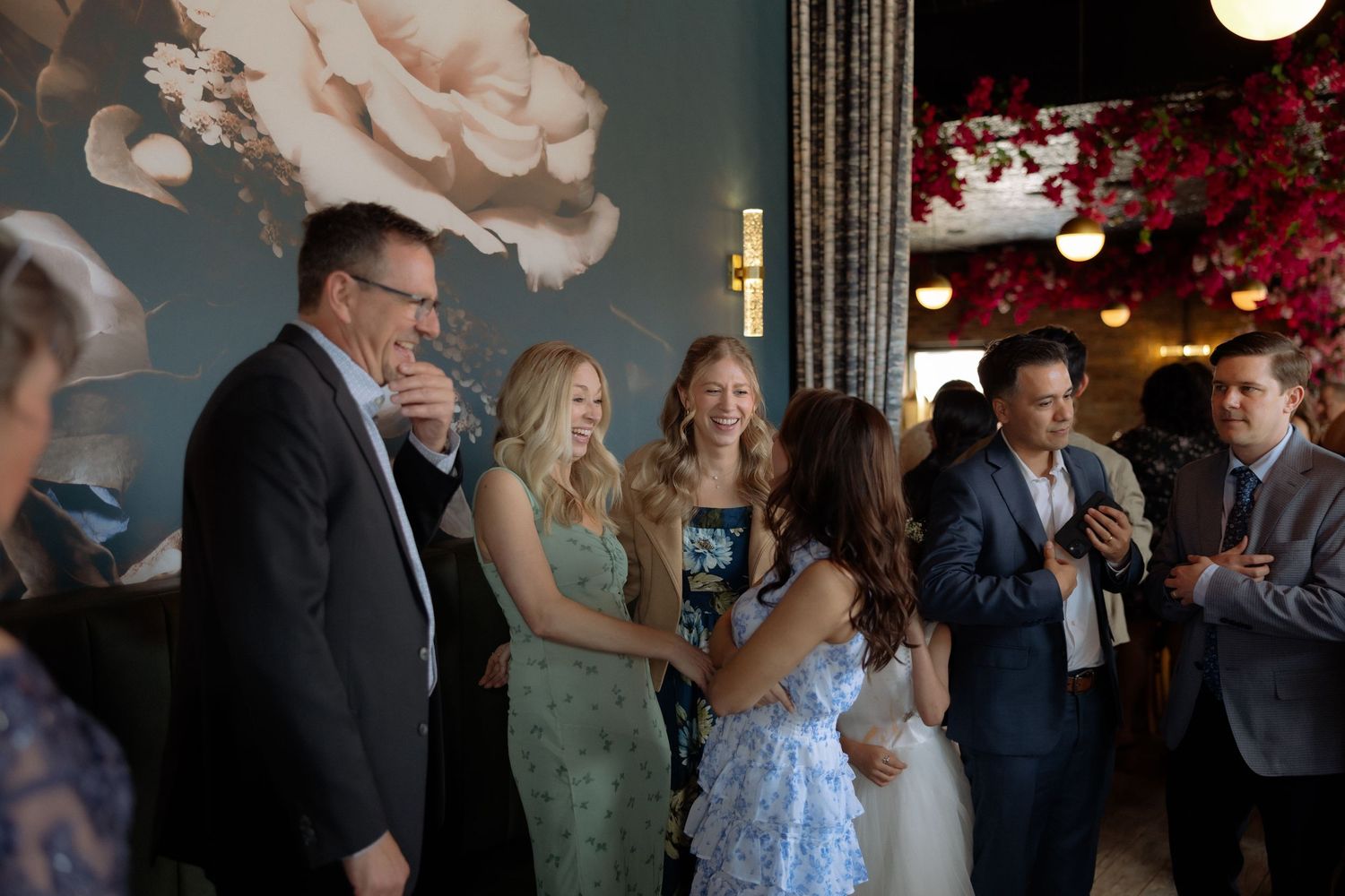 Elegantly dressed guests socializing and laughing at an upscale indoor event with floral wall decor.