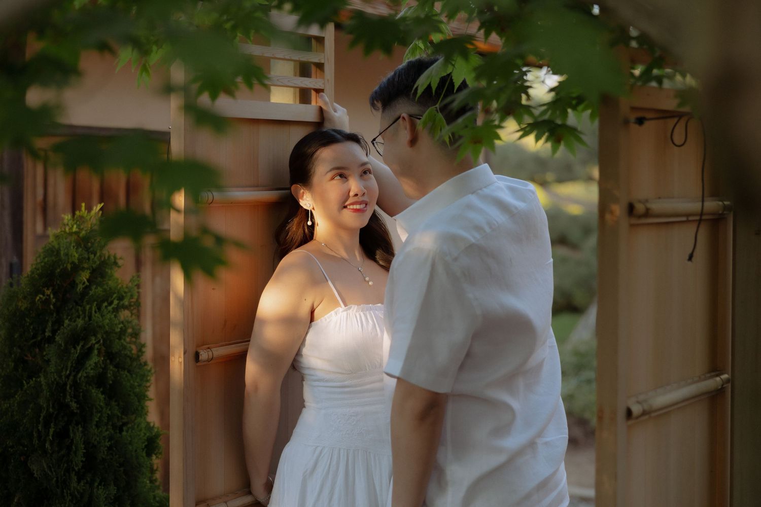 A couple in white outfits share a romantic moment in a Japanese garden setting with lush green foliage.