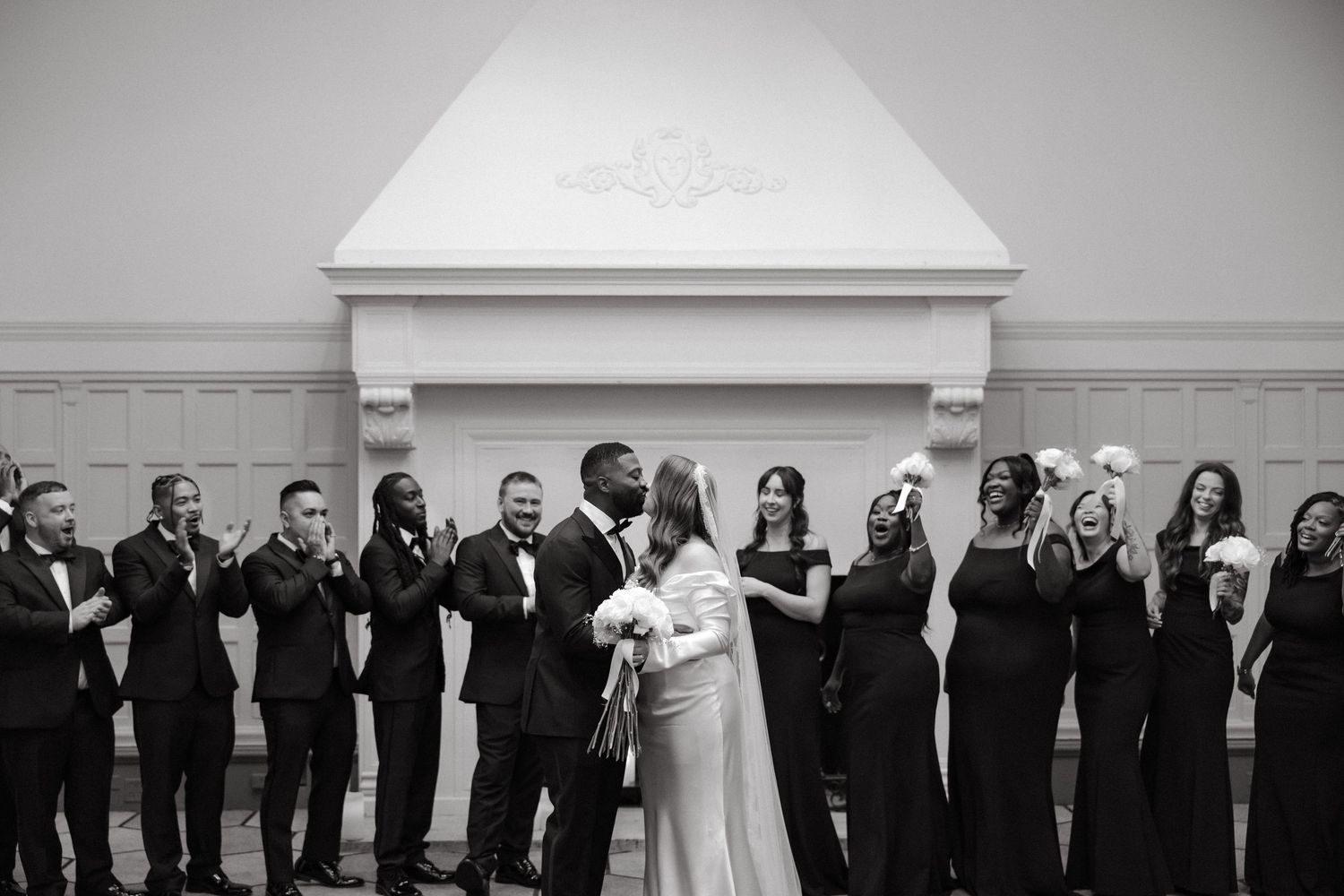 Black and white wedding photo of couple kissing at altar surrounded by applauding bridal party in elegant venue.