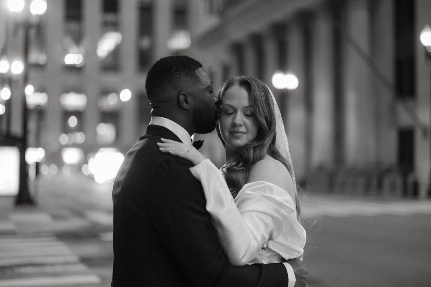 Black and white photo of an interracial couple embracing on a city street at night, bride in white gown.
