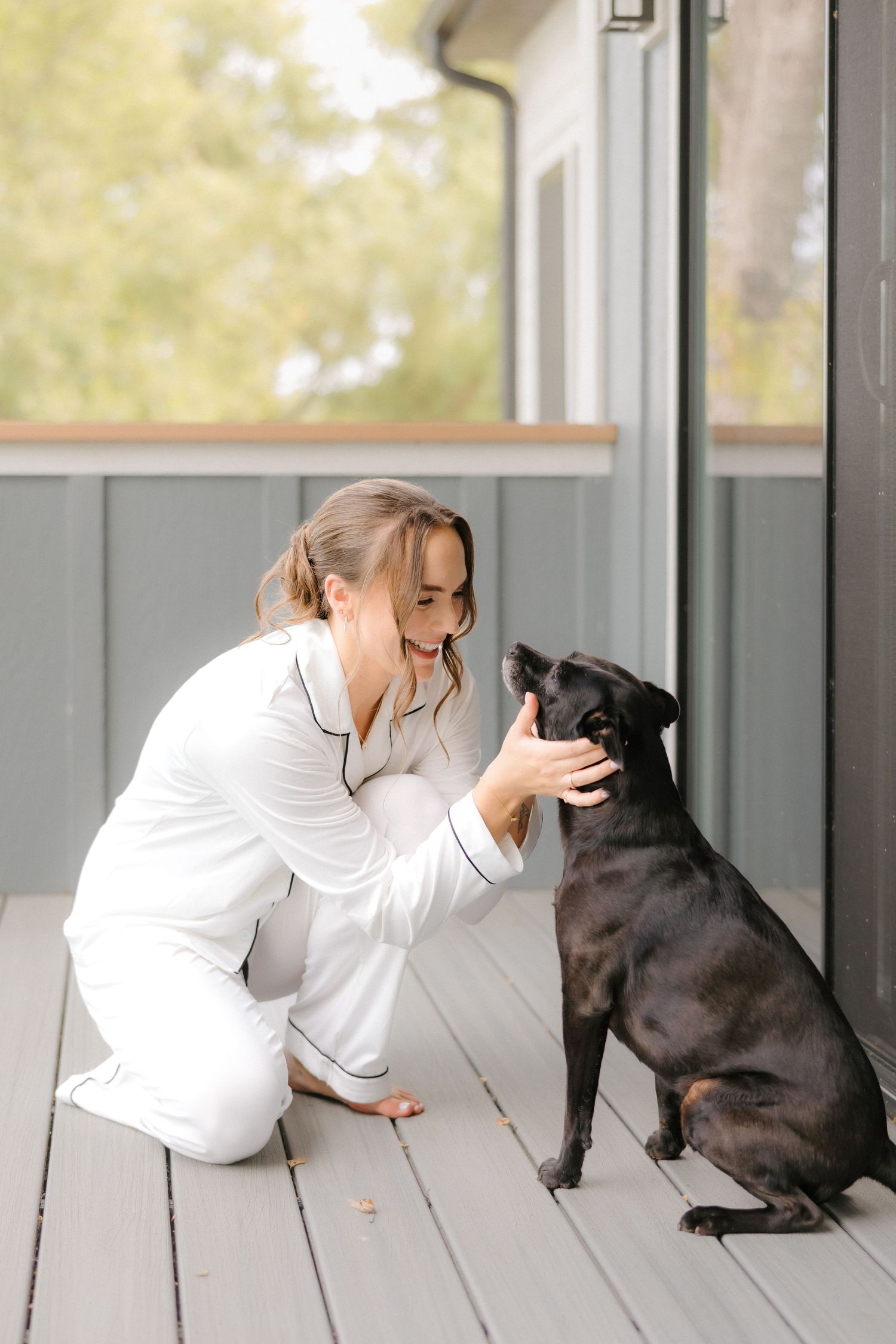 Bride in white pajamas crouches on gray porch deck petting a small black dog on wedding morning.