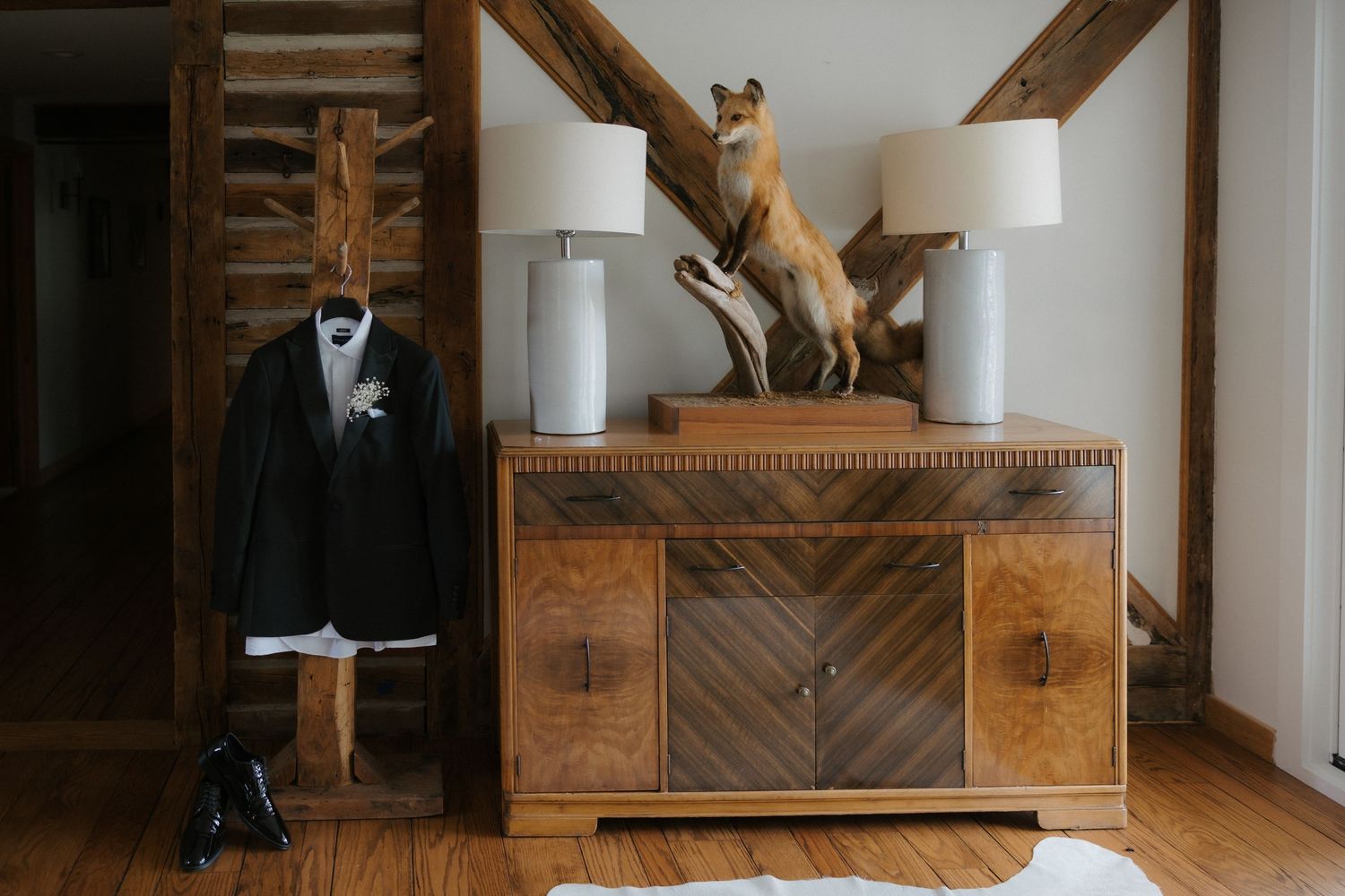 Groom's black suit on hanger beside rustic dresser with fox taxidermy and lamps.