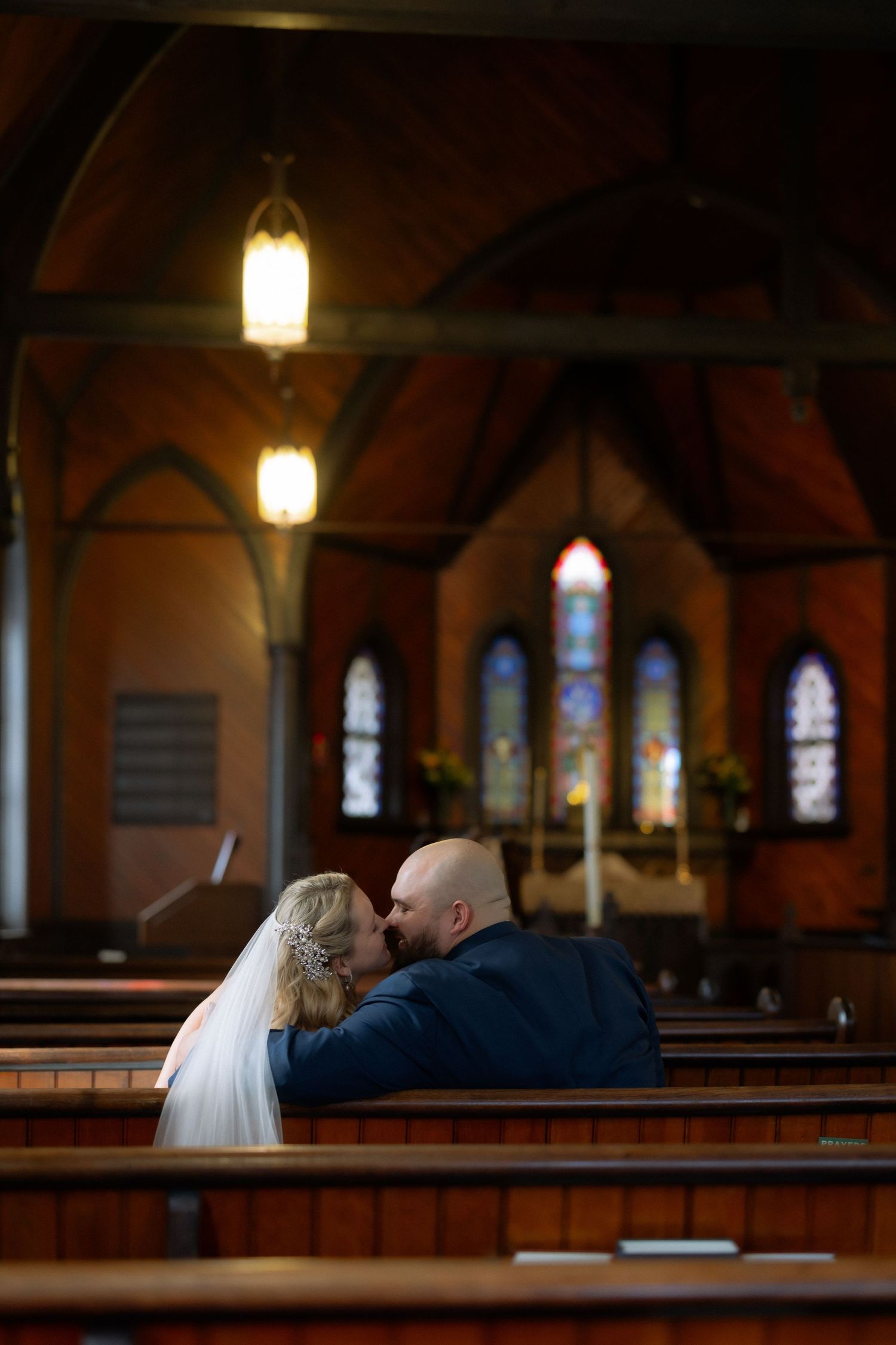 Bride and groom share a kiss in a wooden church pew with stained glass windows and warm lighting in the background.