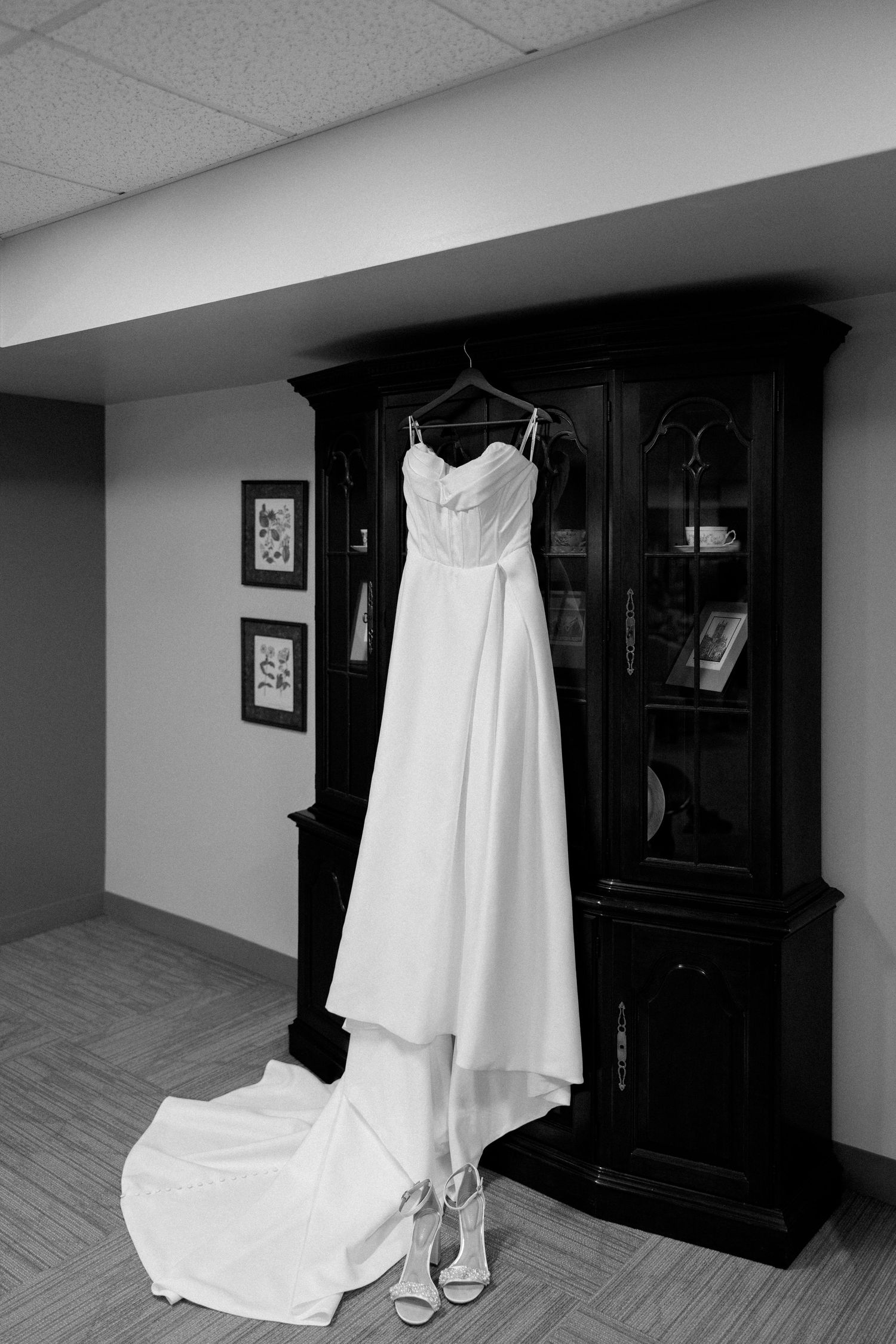 Black and white photo of an elegant wedding gown hanging on a dark wood cabinet with bridal heels below.