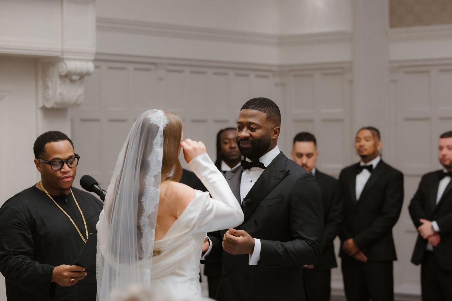 Groom lifts bride's veil during elegant wedding ceremony, officiant and groomsmen in tuxedos stand nearby.