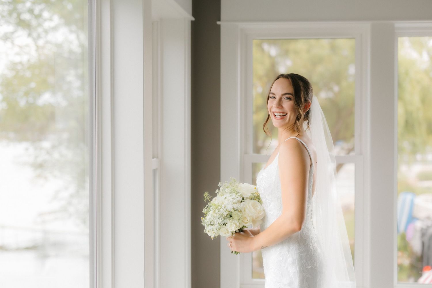 Radiant bride in white lace gown holding white floral bouquet, smiling softly beside bright natural light windows.