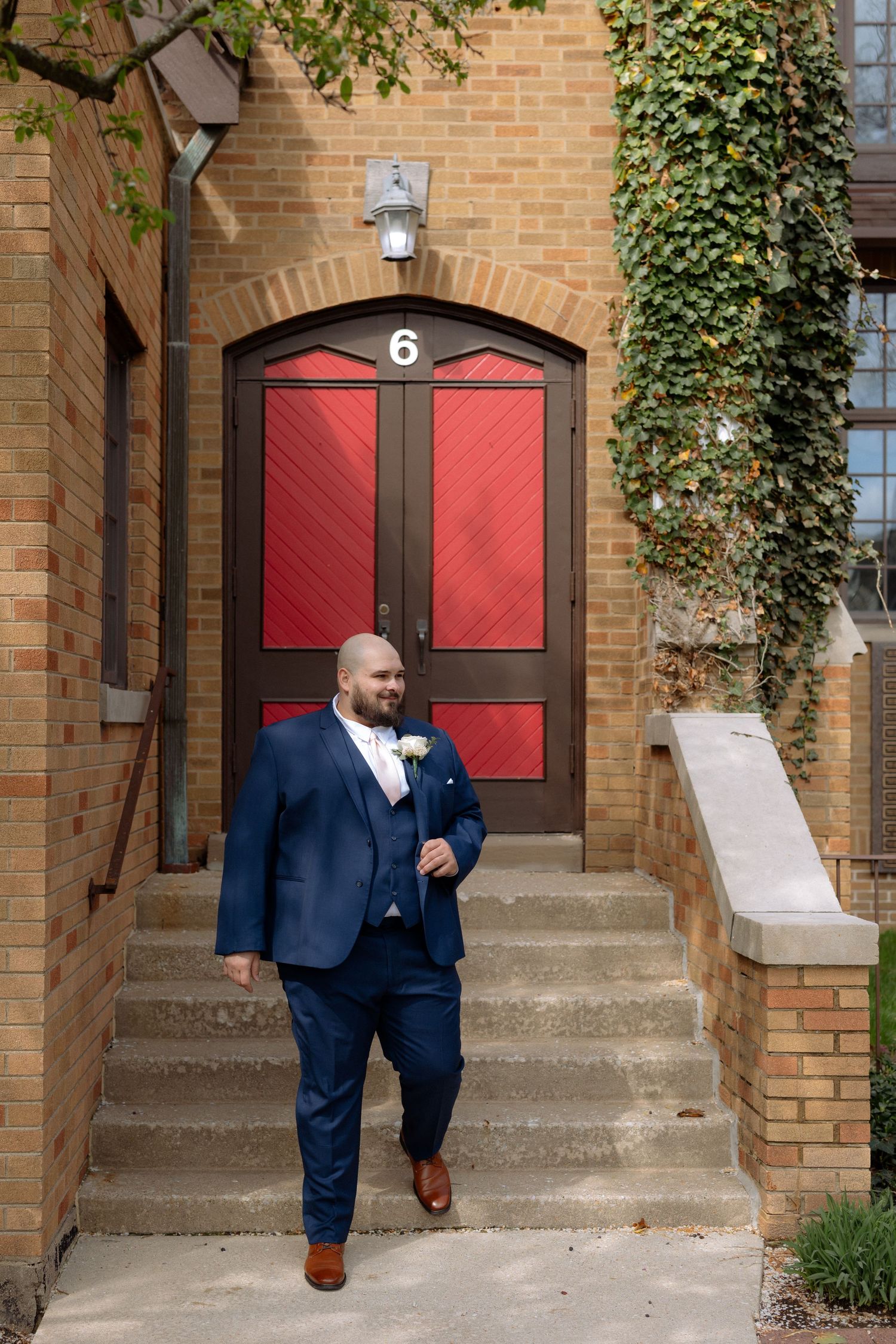 Groom in navy blue suit with boutonniere walks down steps in front of red double doors marked with number 6.