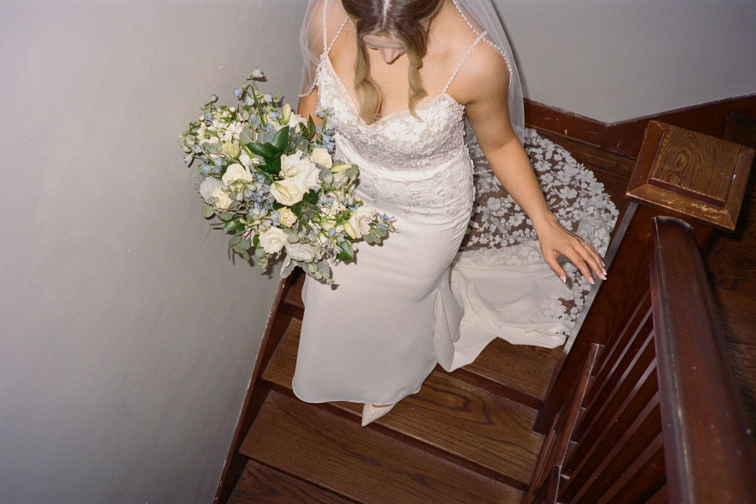 Aerial view of bride in lace wedding gown holding white bouquet descending wooden staircase.