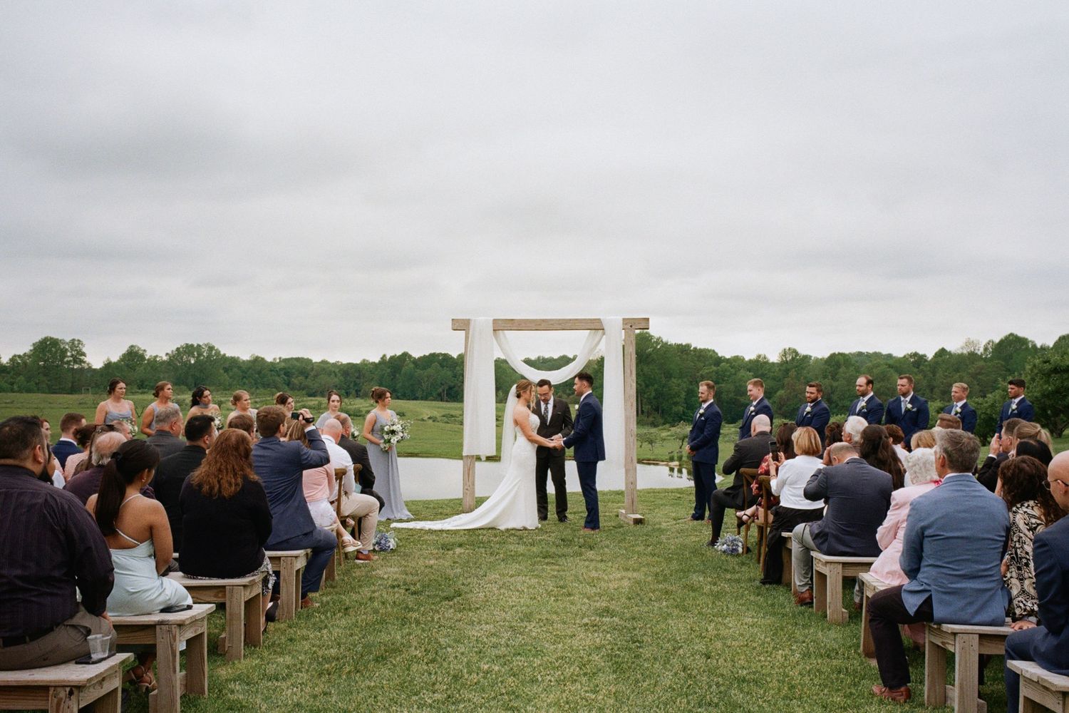 Outdoor wedding ceremony on green lawn with wooden arch, bride and groom at altar, guests seated on rustic benches under cloudy sky.