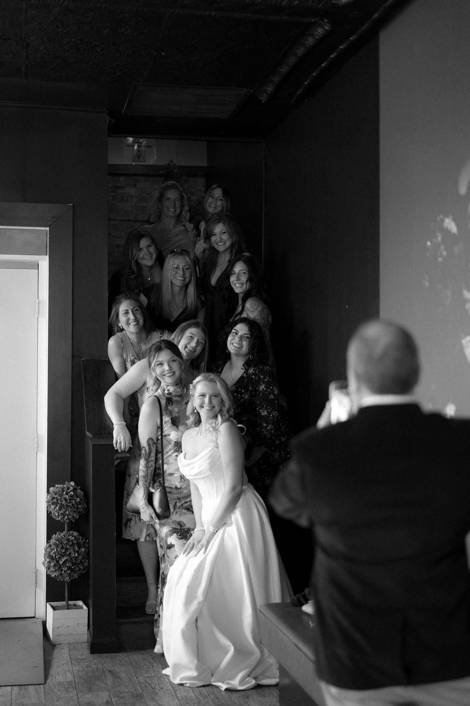 Black and white photo of a bride posing with bridesmaids while a man photographs the group at a wedding venue.